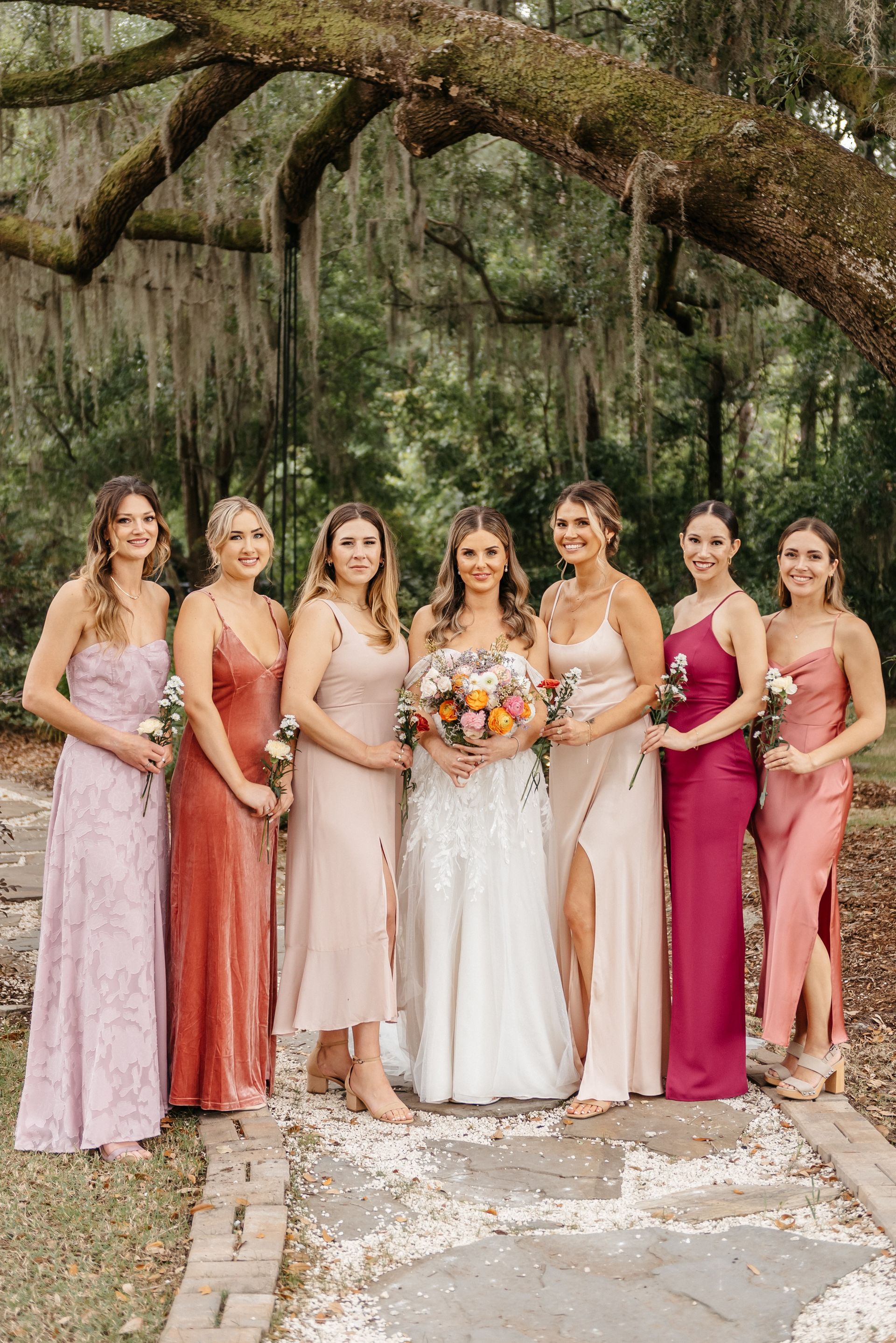 Bridesmaids and bride posing under a large oak tree at an outdoor wedding, holding a bouquet.