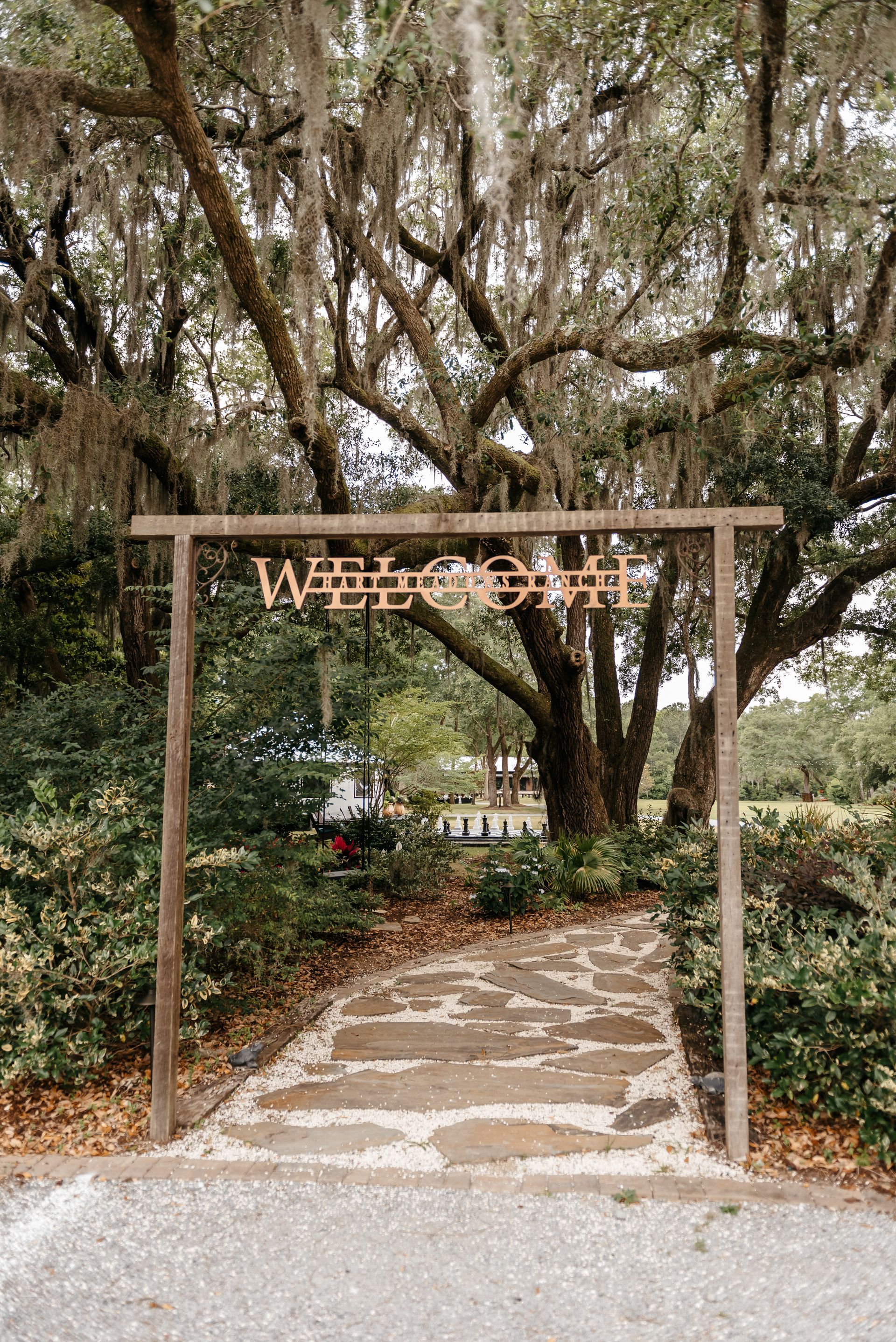 Wooden welcome arch over a stone path beneath large trees at a garden entrance