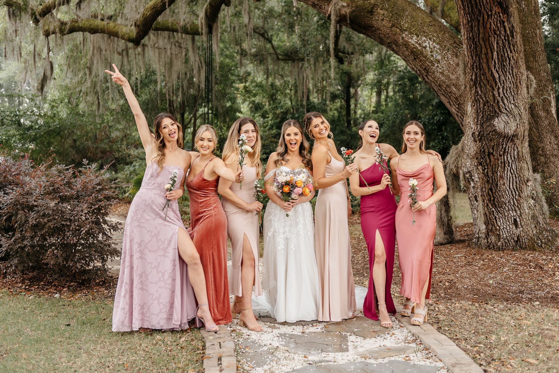 Six women posing outdoors in formal dresses at a wedding, one holding a bouquet under a large tree