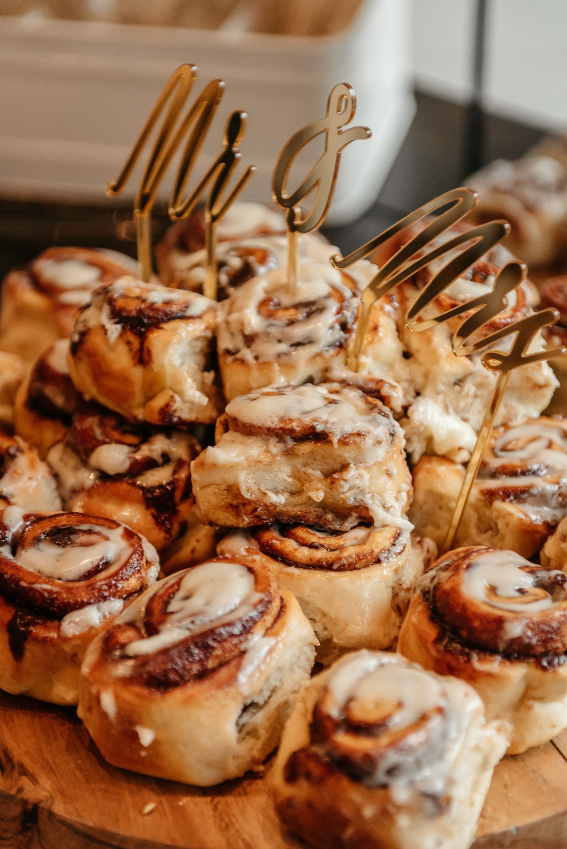 Iced cinnamon rolls on a wooden tray with a gold “Mrs” topper for a wedding dessert display