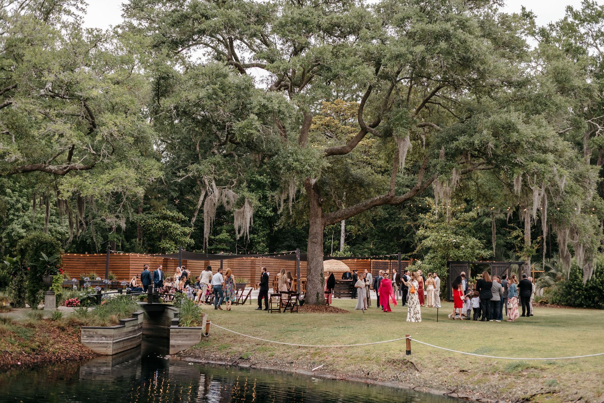 People gathered in a park under large trees, with a pond in the foreground and a brick wall behind.