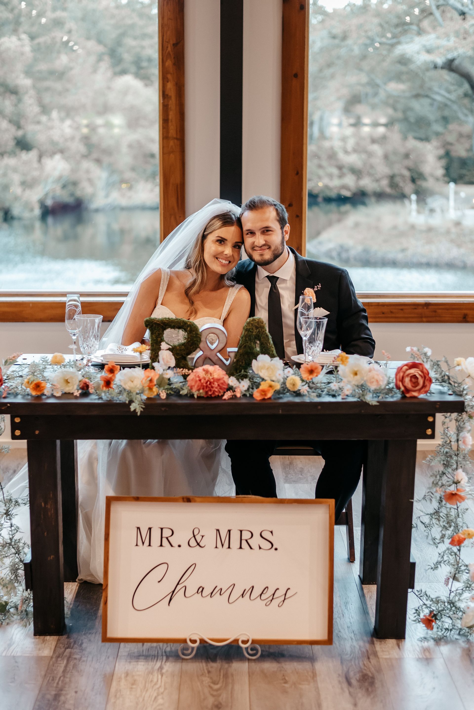 Newlyweds pose at a decorated wedding table with a “Mr. & Mrs.” sign and autumn flowers.