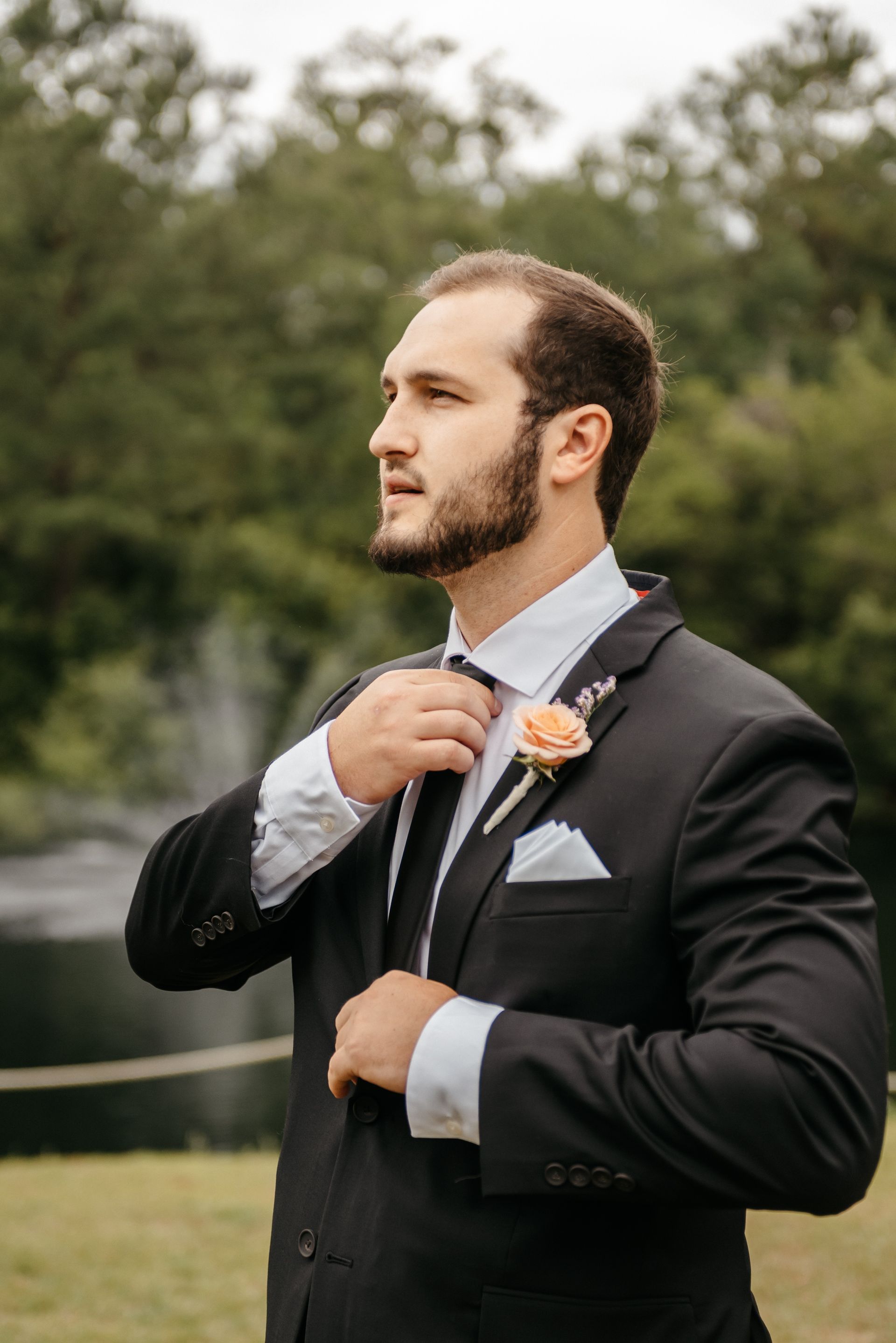 Man in a black suit adjusting his tie outdoors, with trees blurred in the background.