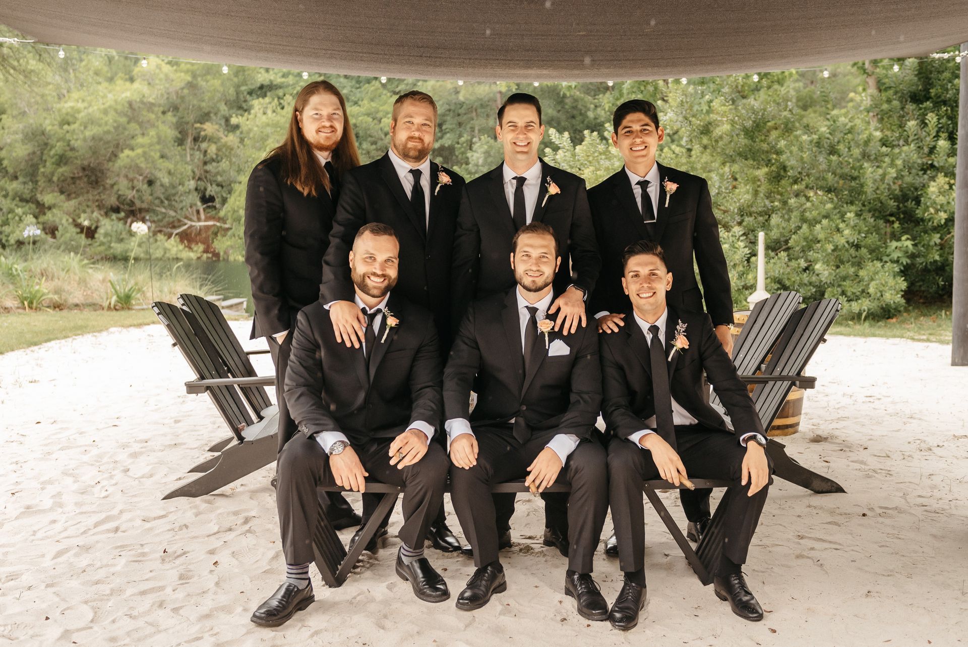 Seven formally dressed people pose outdoors under a pavilion, with trees and benches in the background.