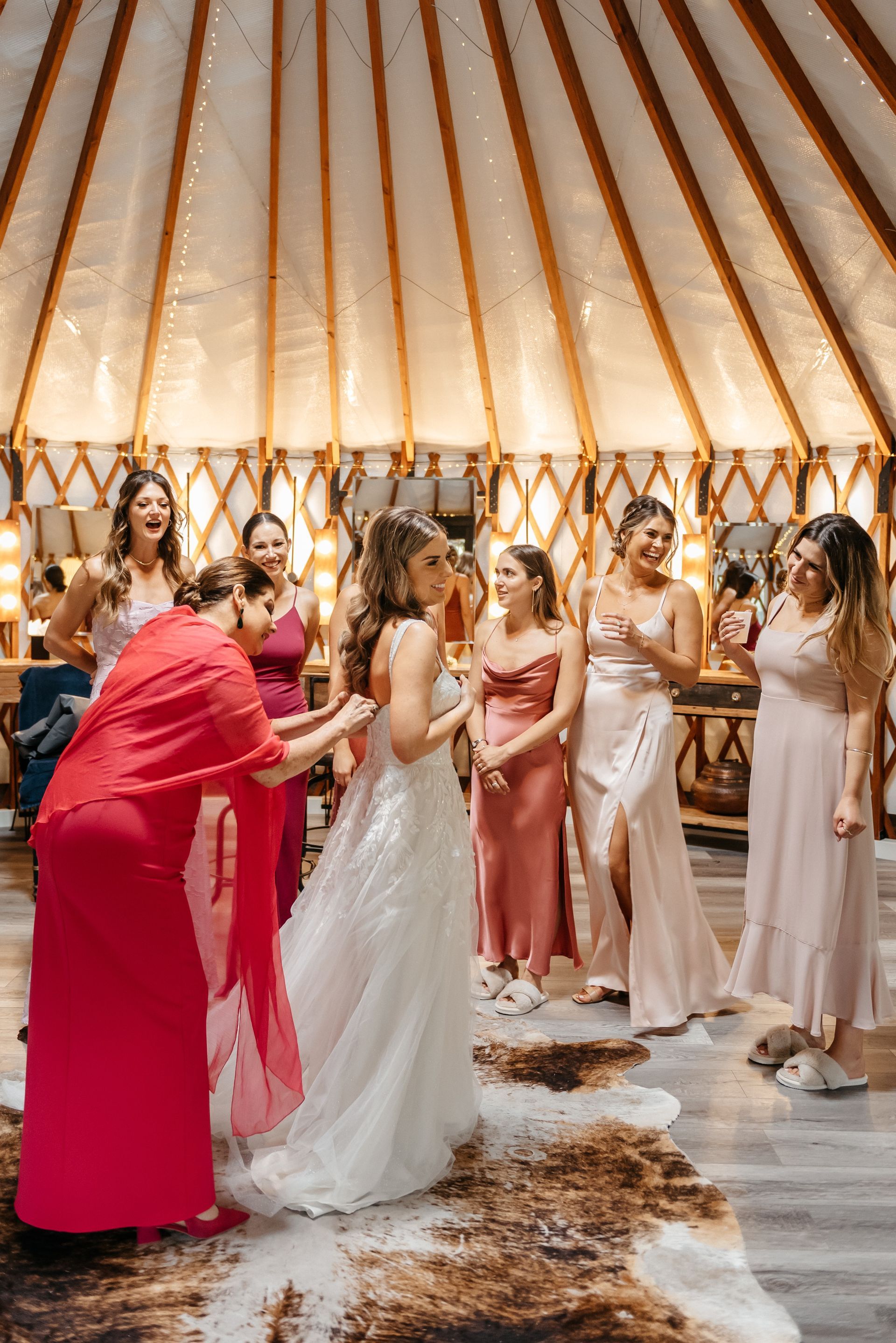 Bridal party at a teepee-style ceremony, with women in white and pink dresses embracing and smiling indoors