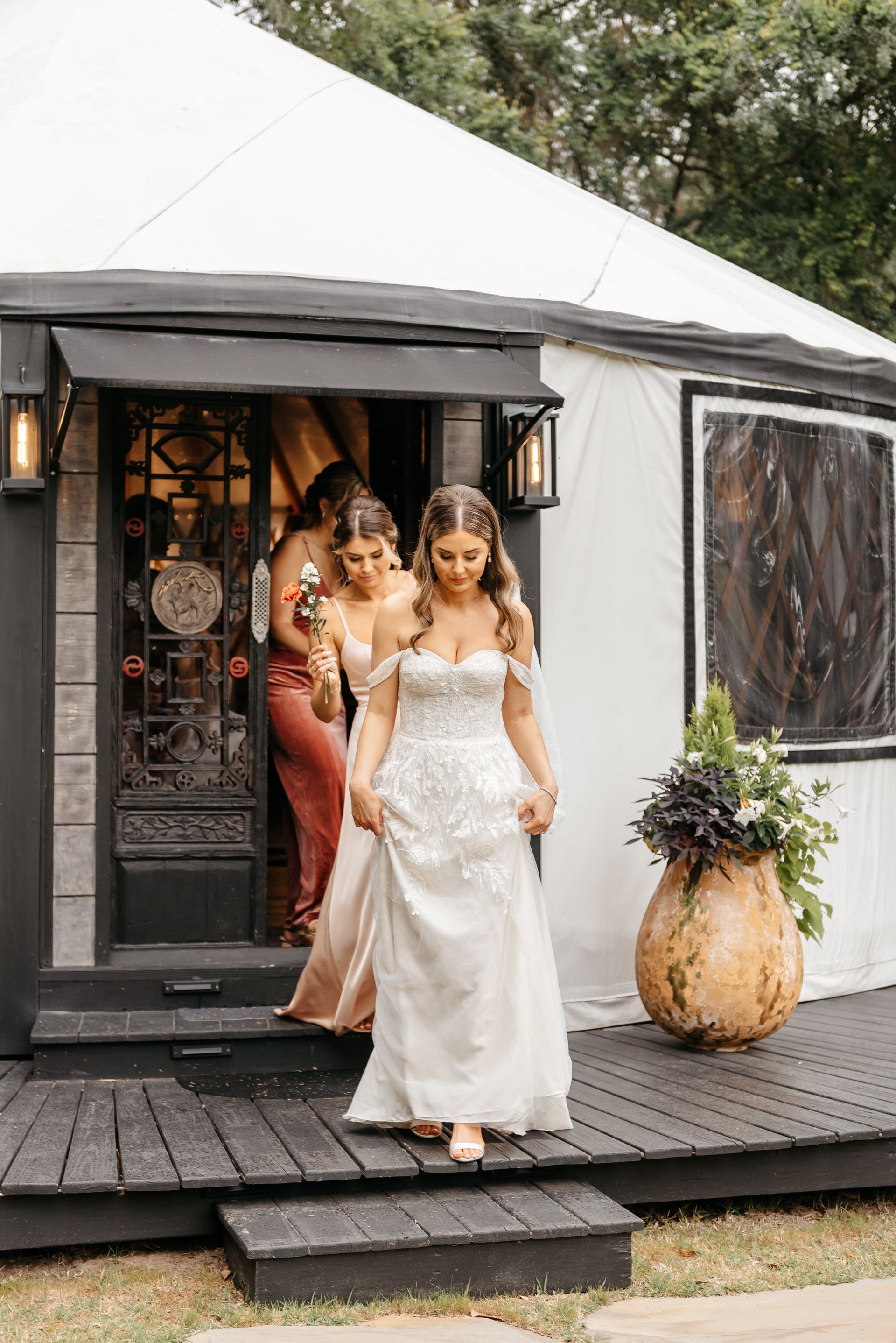 Bride in white dress stepping from a tented venue, with bridesmaid behind her and floral décor by the entrance