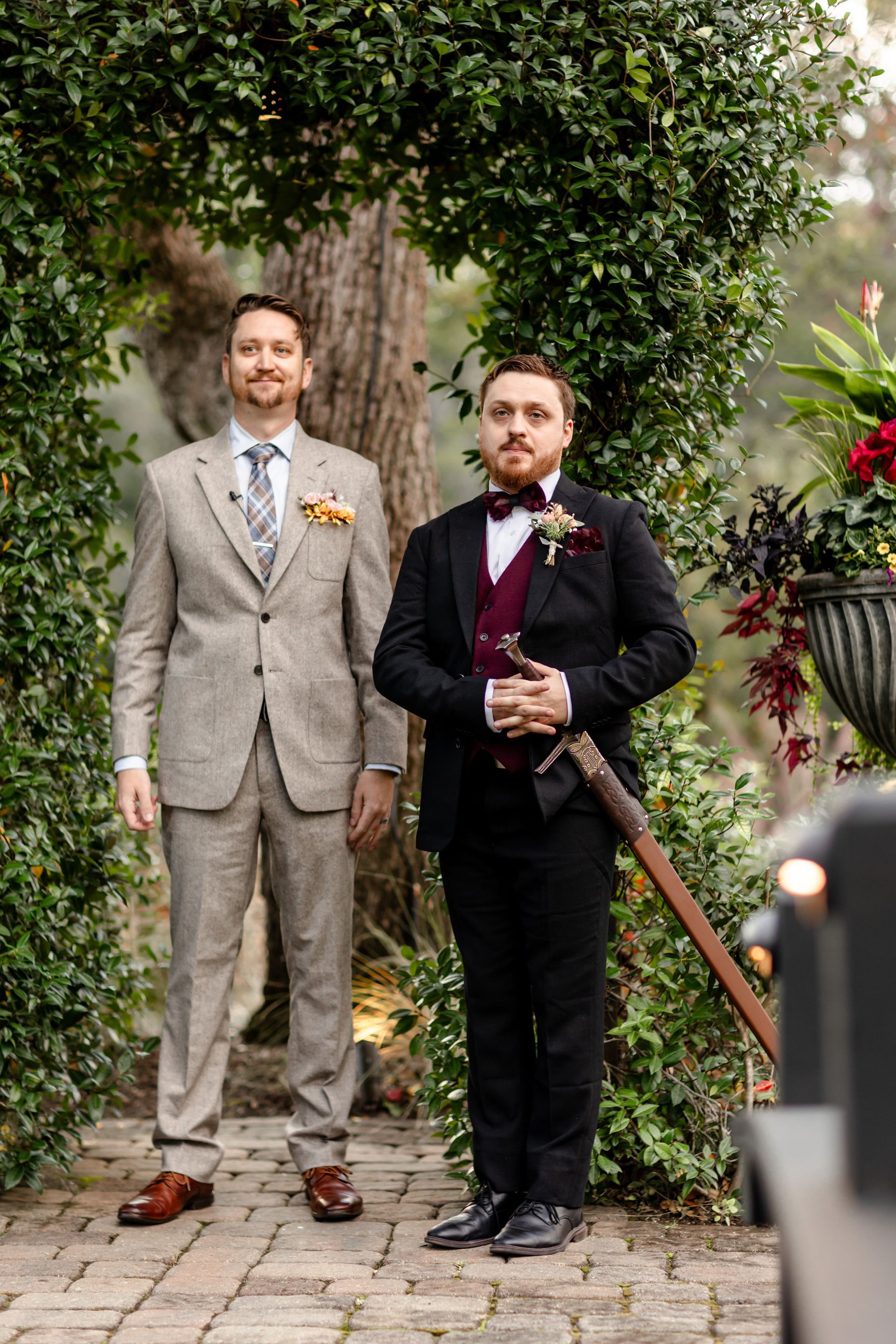 Two men in formal suits standing outdoors under a tree at a wedding venue.