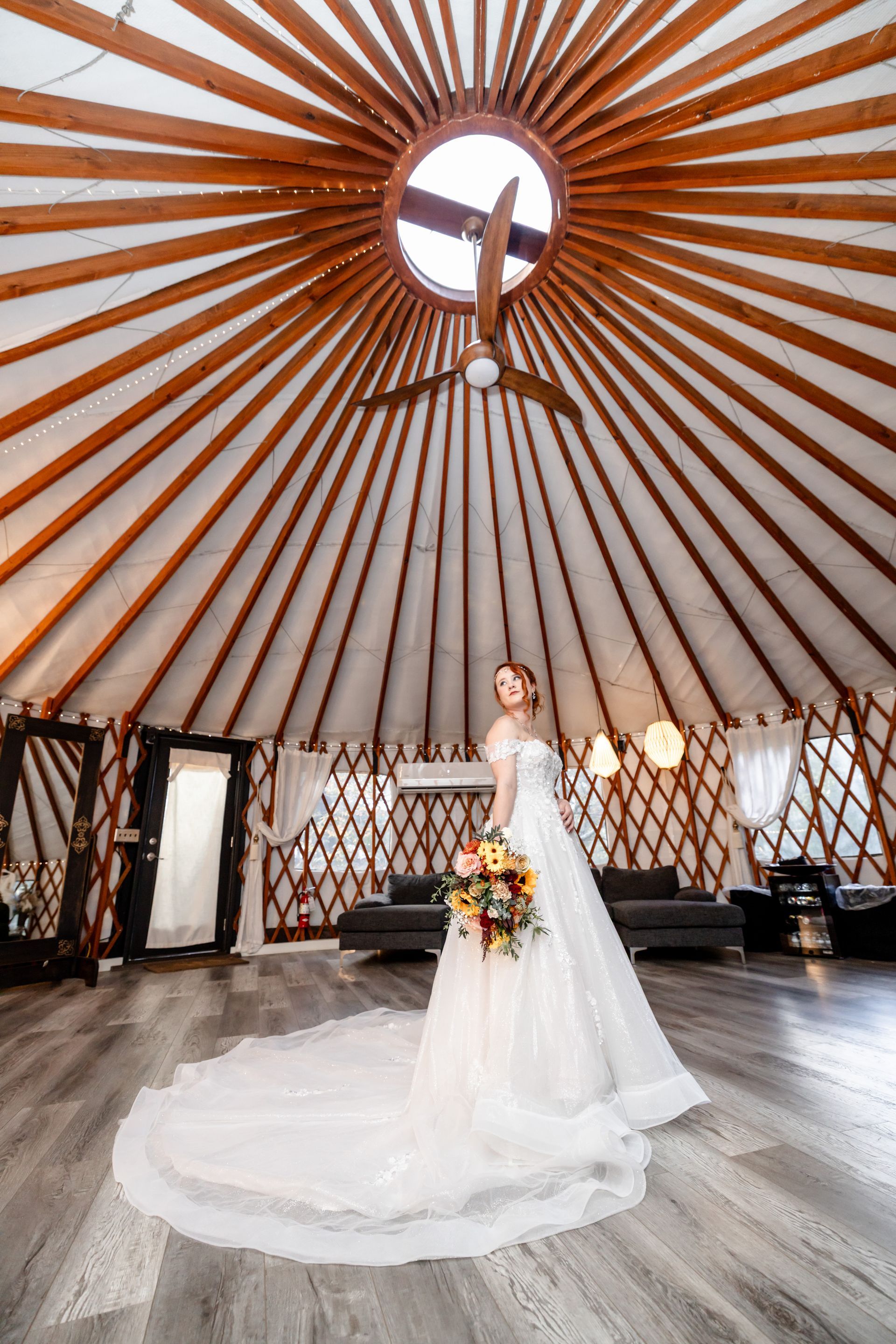 Bride in a white gown holding a bouquet inside a circular wooden yurt with a skylight above