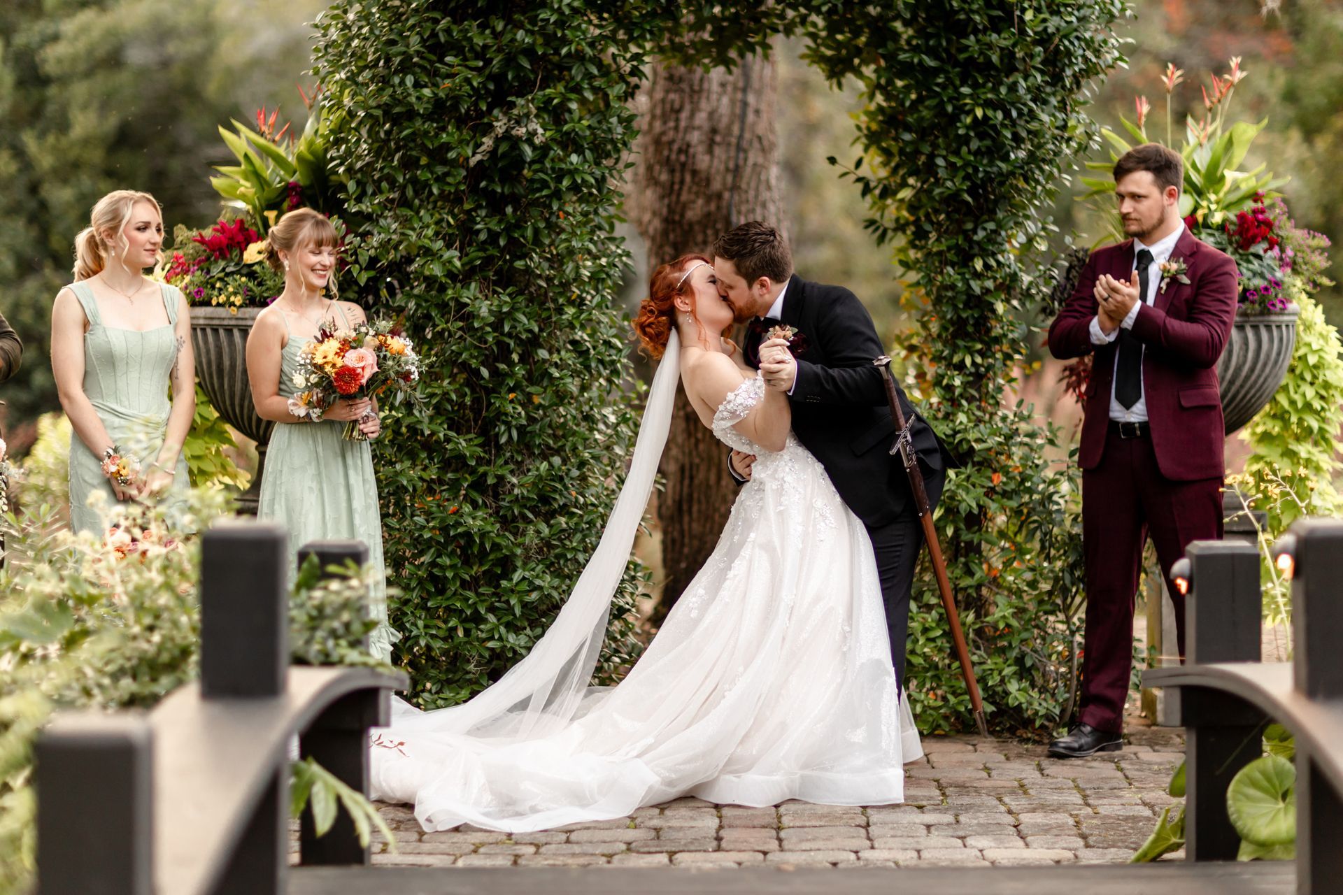 Wedding couple kissing under an ivy arch, with attendants standing nearby in a garden.
