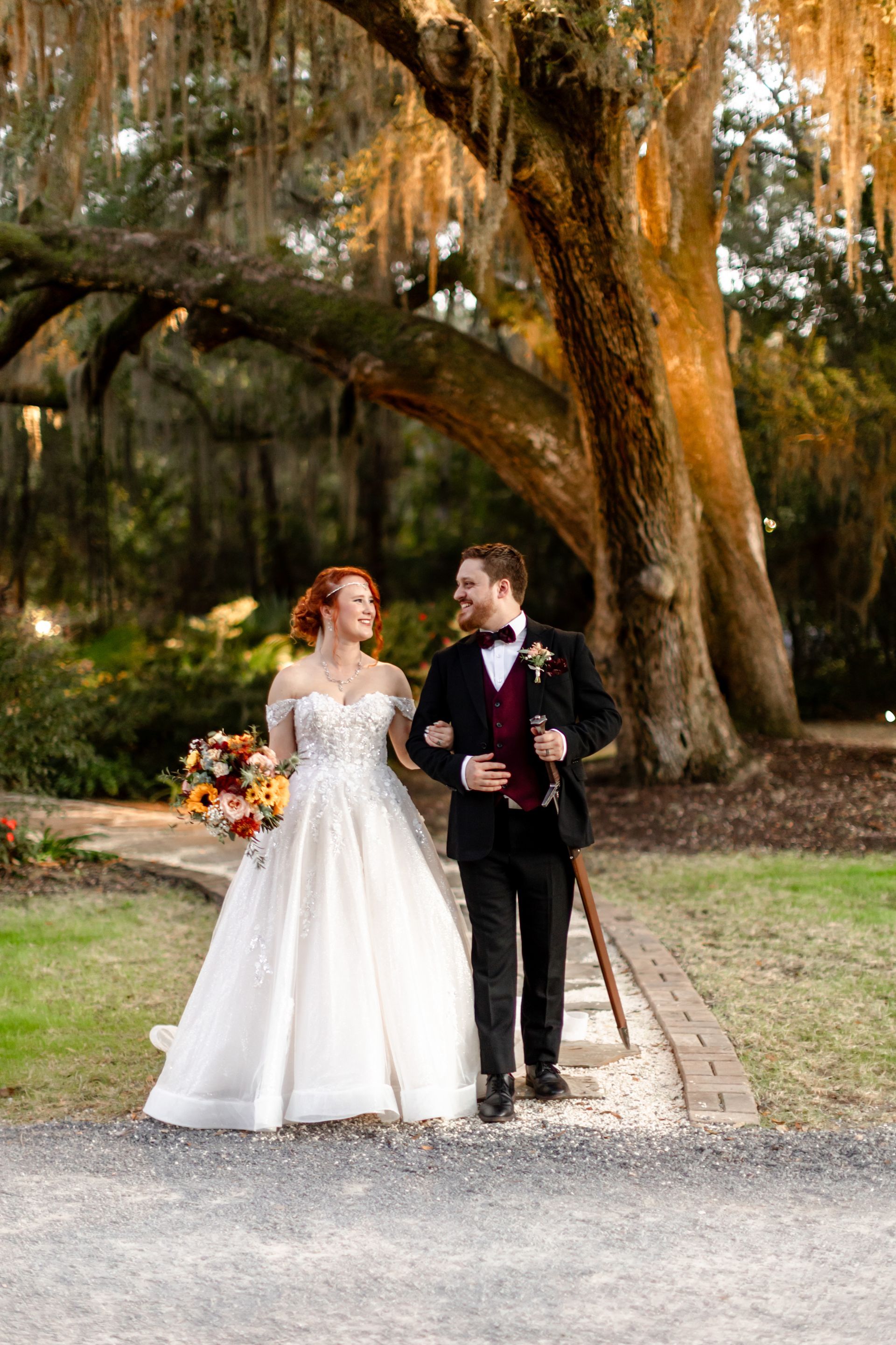 Bride in a white gown and groom in a black suit walking arm in arm under large oak trees.