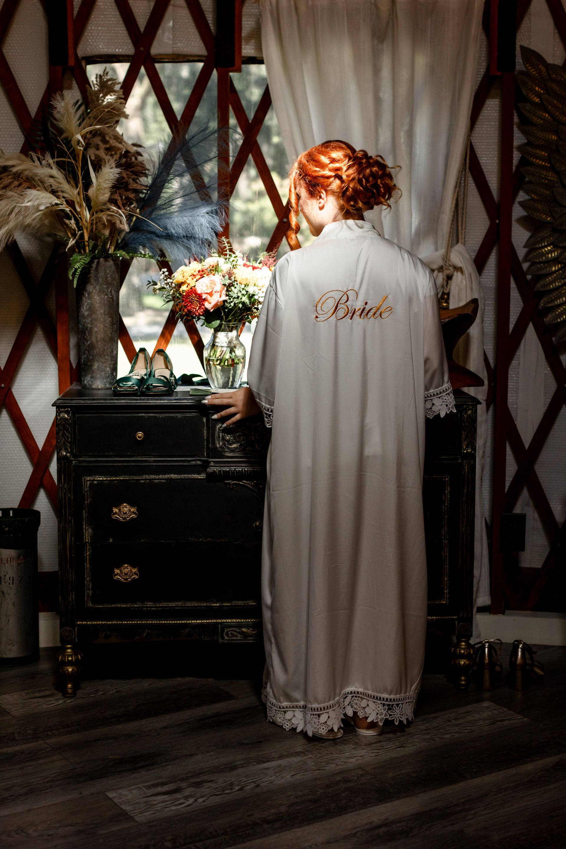 Person in a white robe standing at a dresser with flowers in a dimly lit room