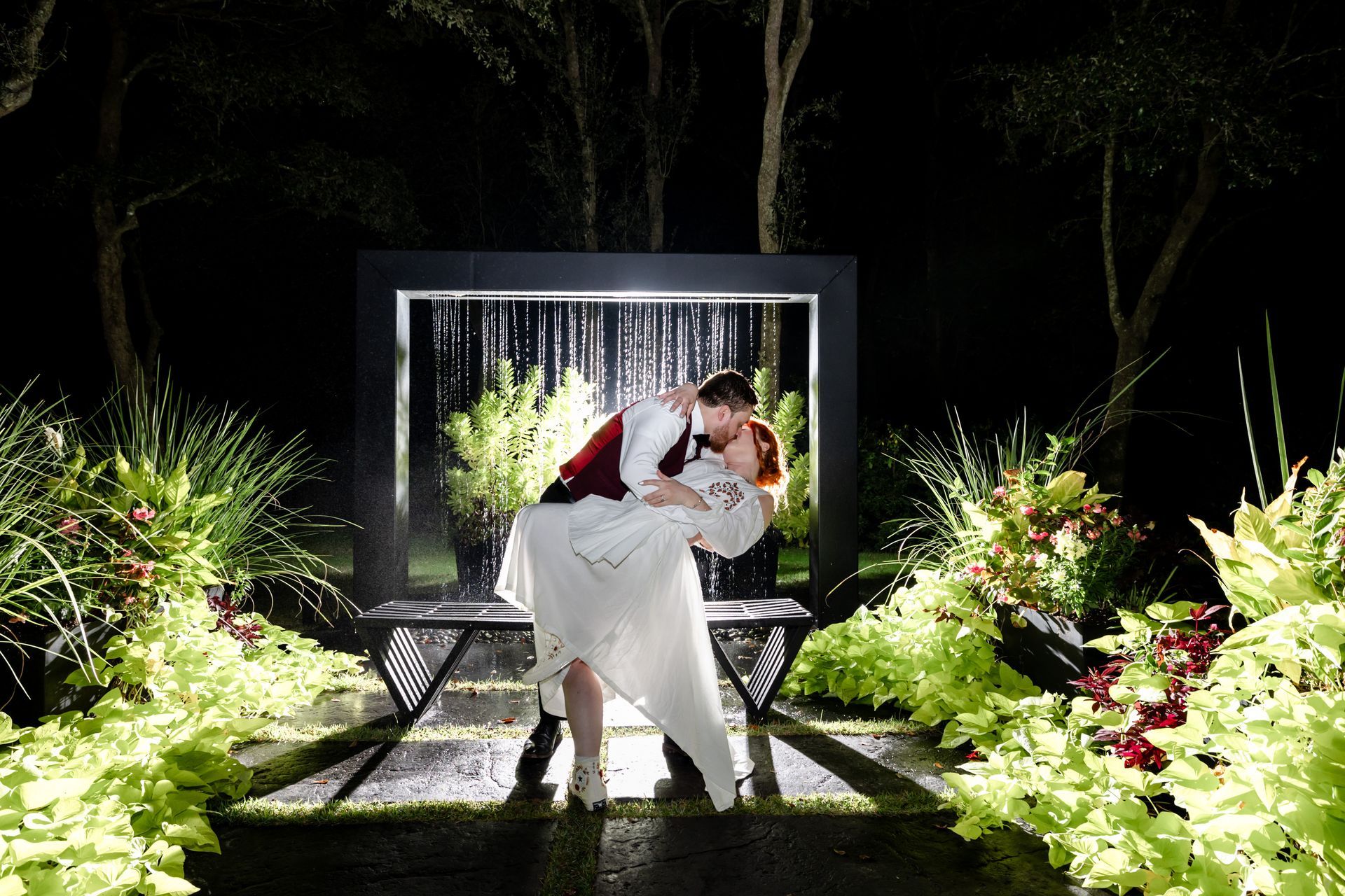 Two people embracing on a lit garden path at night, surrounded by greenery and white flowers