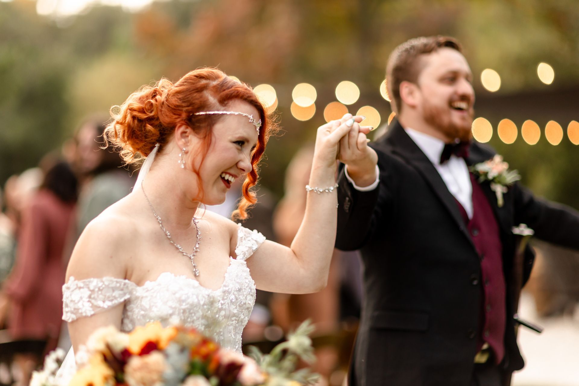 Smiling couple holding hands and dancing at an outdoor wedding, with blurred lights in the background