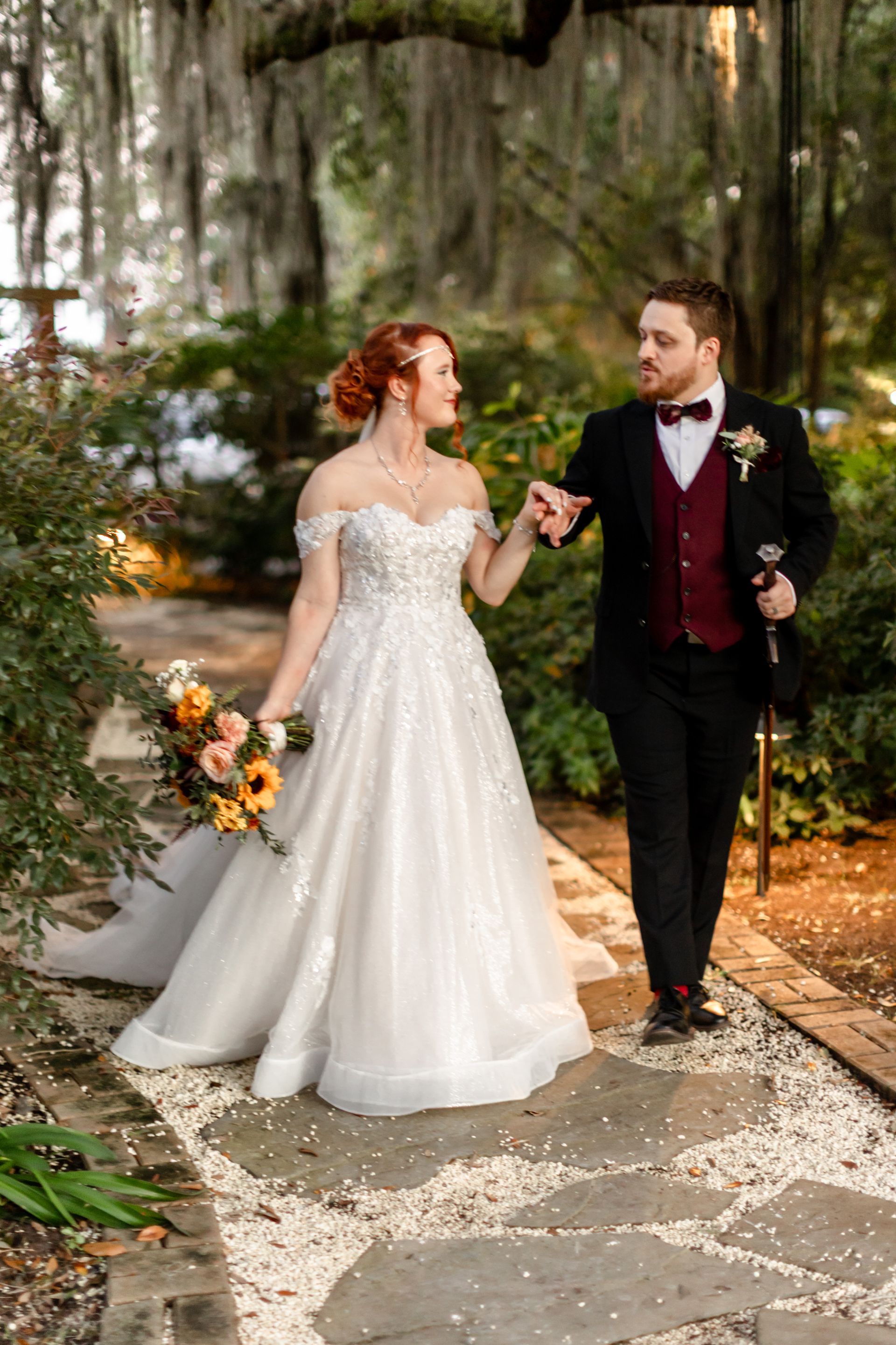 Bride and groom walking hand in hand through a wooded path, bride in white gown, groom in dark suit.
