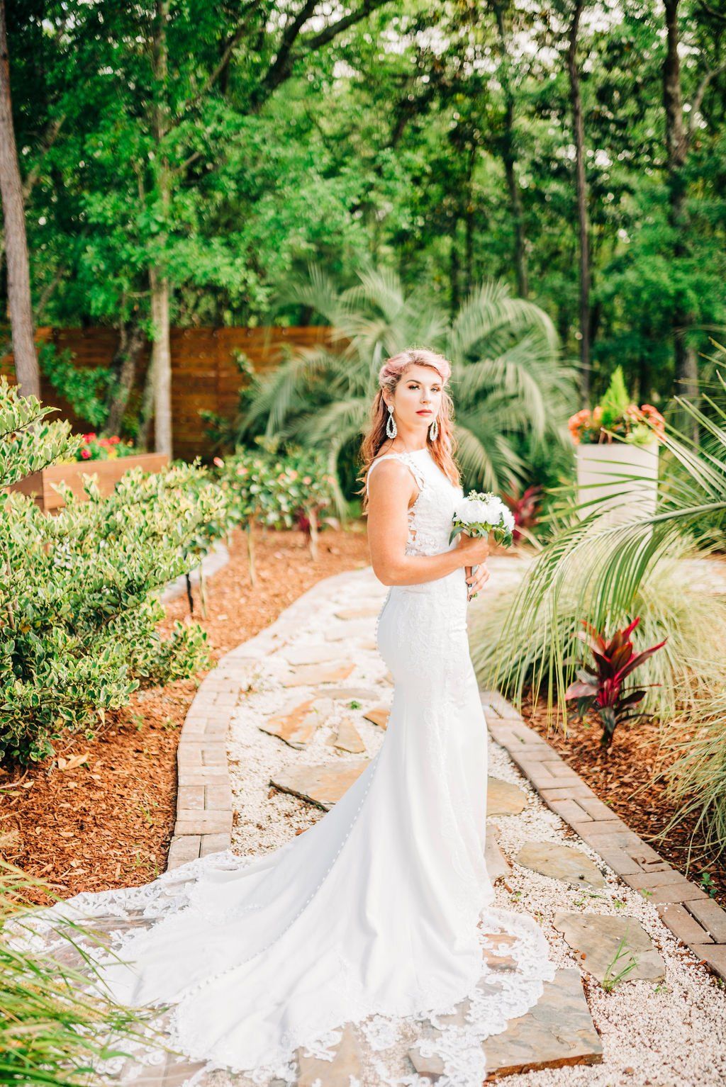 Bride in a white gown standing on a garden path with lush greenery and flowers