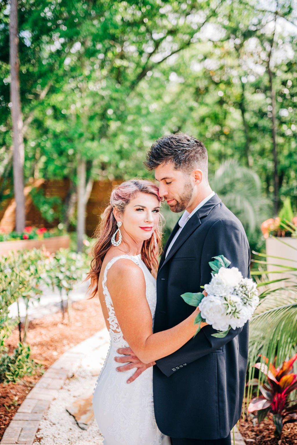 Bride and groom posing in a garden, holding white flowers and smiling by a curved path.