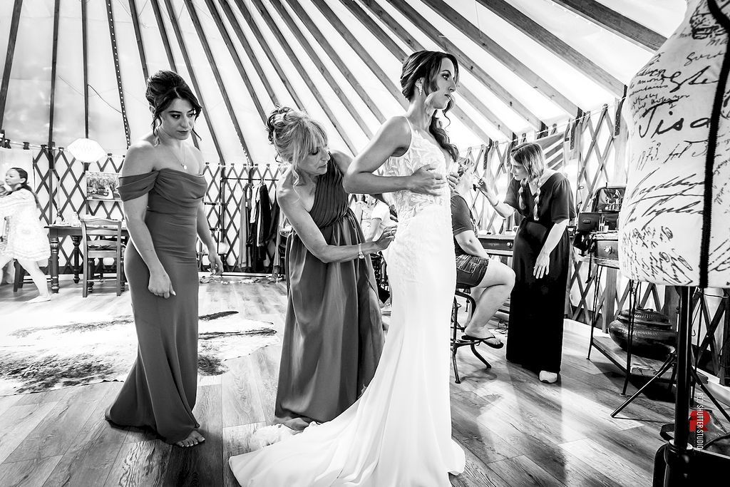Bridesmaids helping a bride adjust her gown inside a striped event tent.