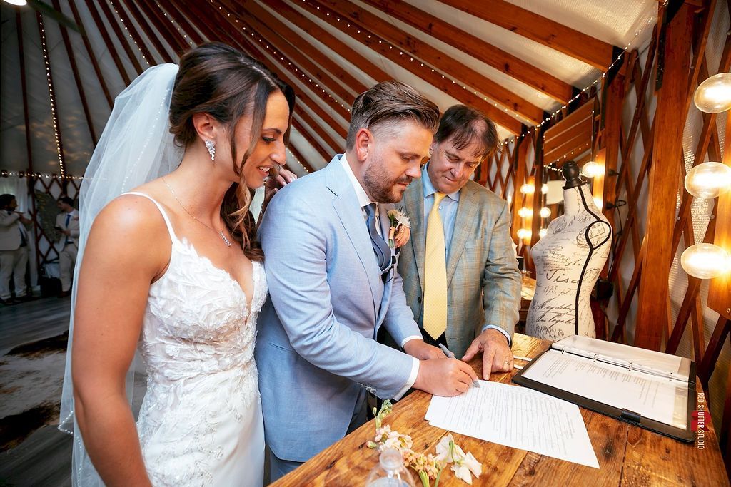 Bride and groom sign a marriage document at a wooden wedding ceremony table.