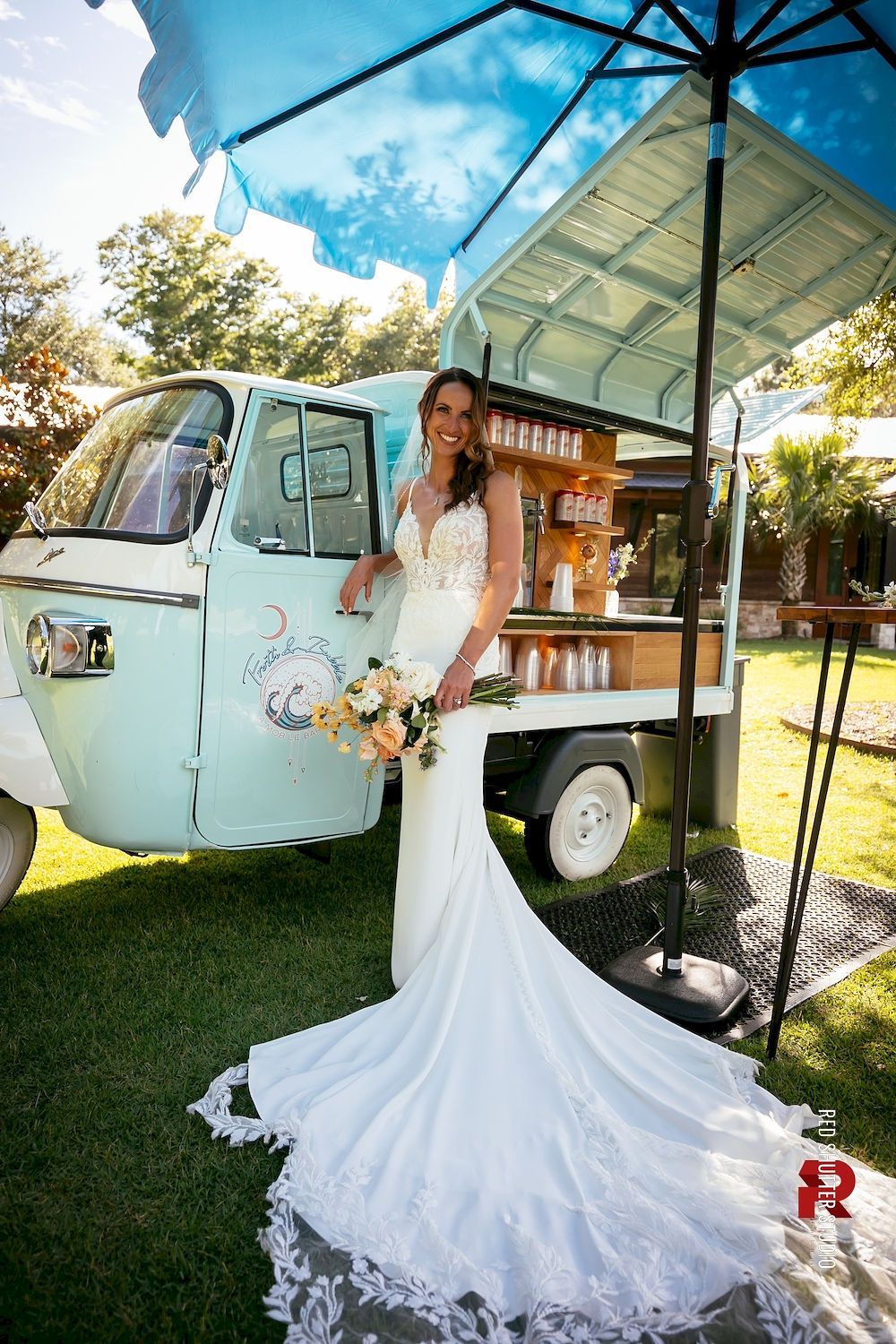 Bride in white gown holding bouquet beside a vintage truck under a blue canopy at an outdoor wedding