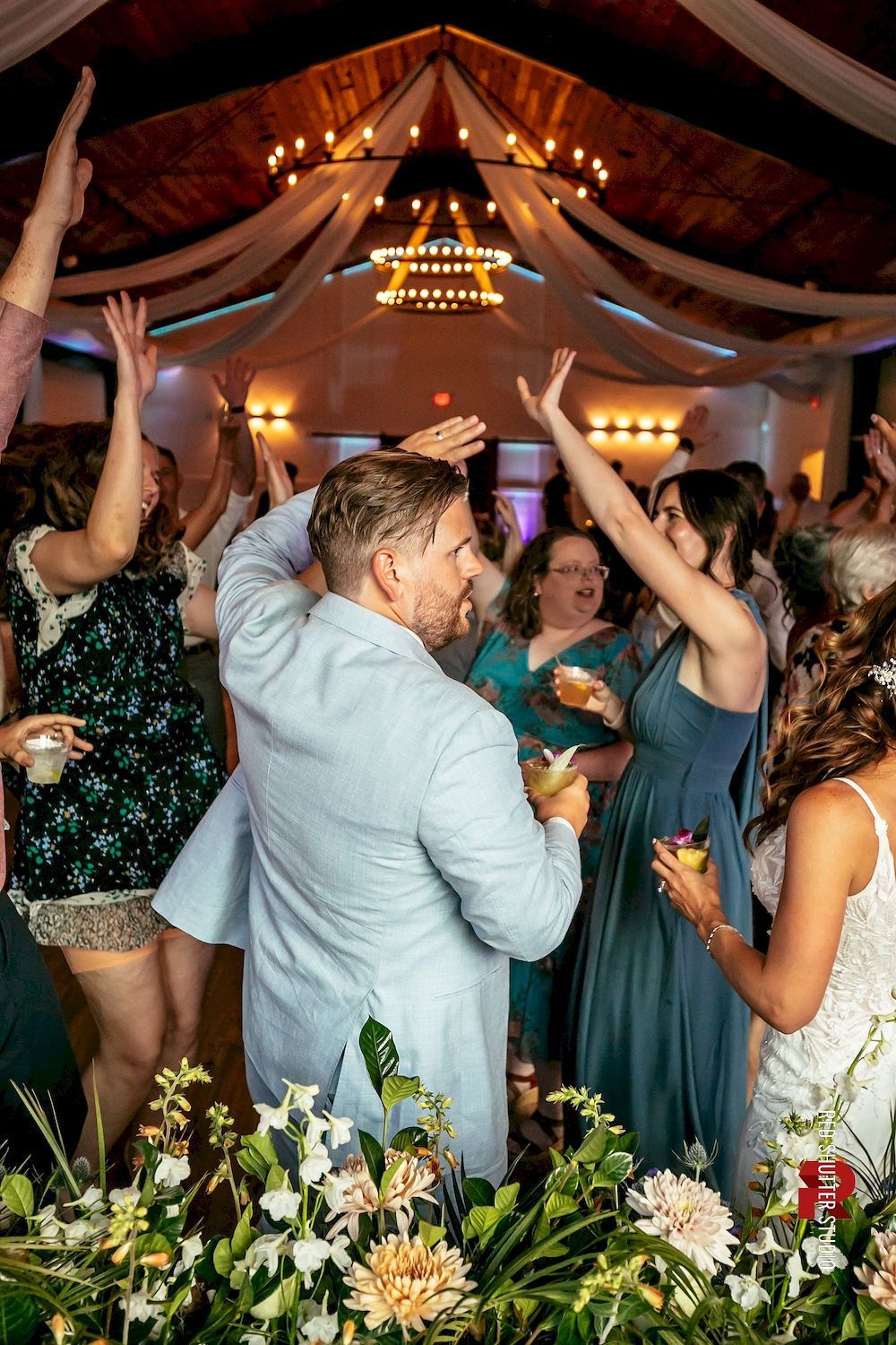 Wedding dance floor under draped lights, guests raising hands around a couple in the center.