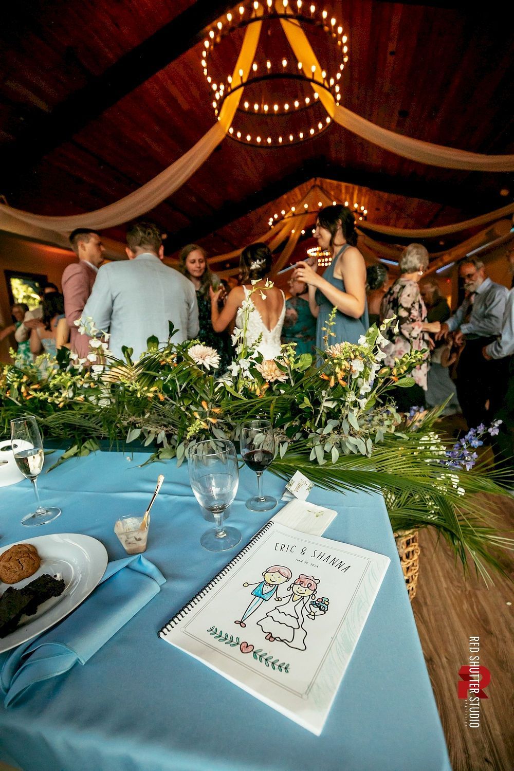 People mingling at a warmly lit indoor reception with long blue tablecloths and floral centerpieces