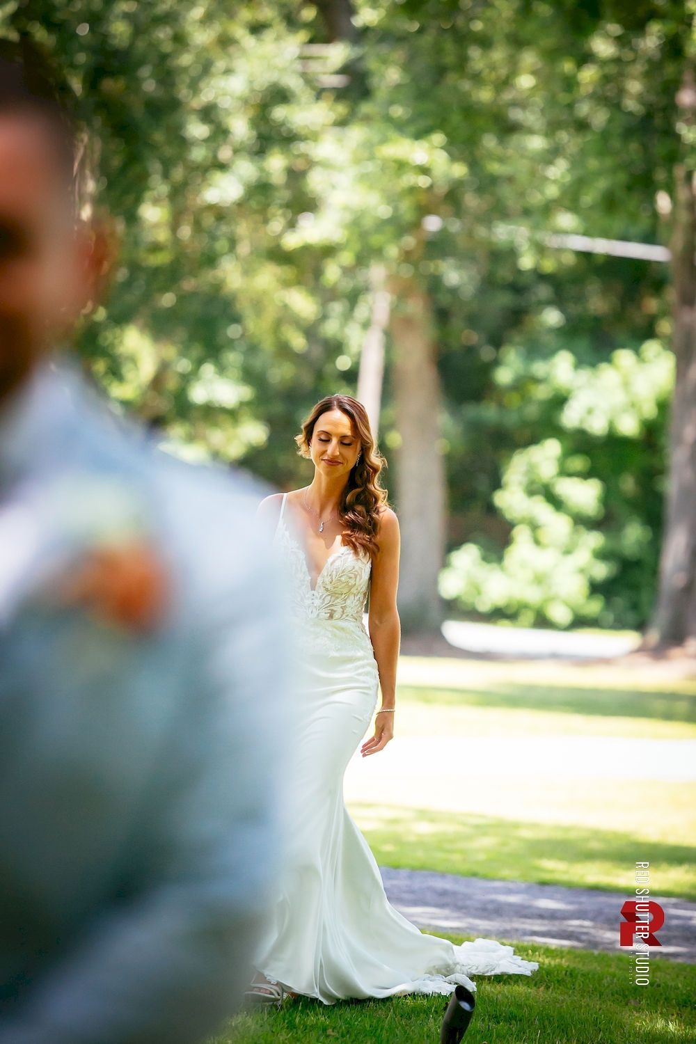 Bride walking outdoors in a white dress, seen between blurred greenery in a sunlit park