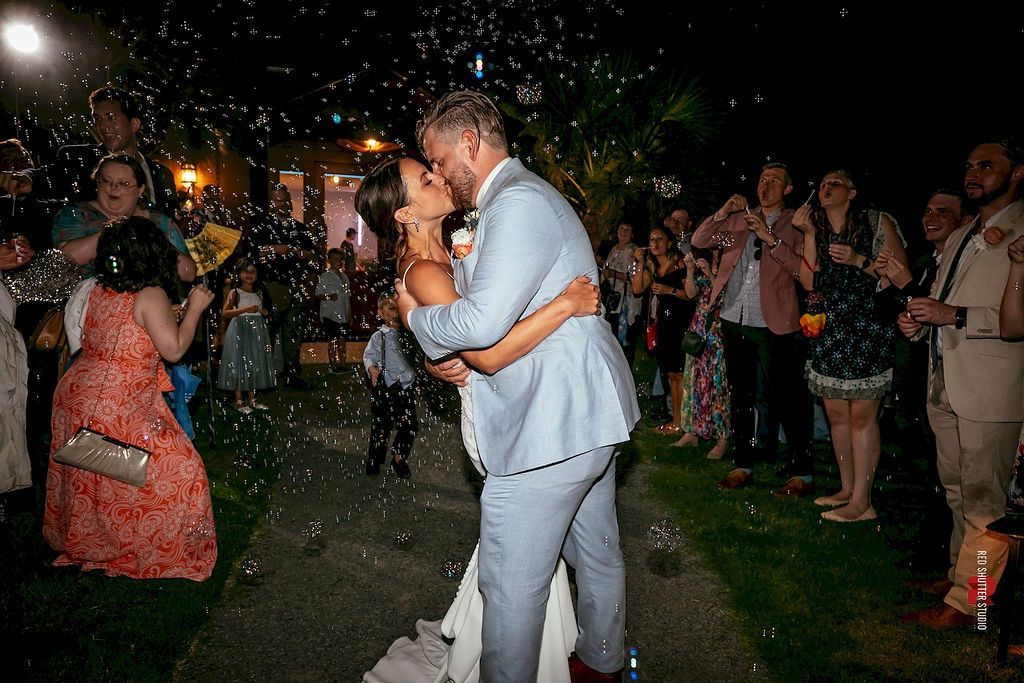 Couple kissing at an outdoor evening celebration, surrounded by guests and confetti.