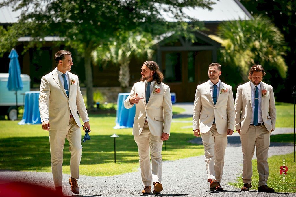 Four people in beige suits walking on a lawn at an outdoor event.