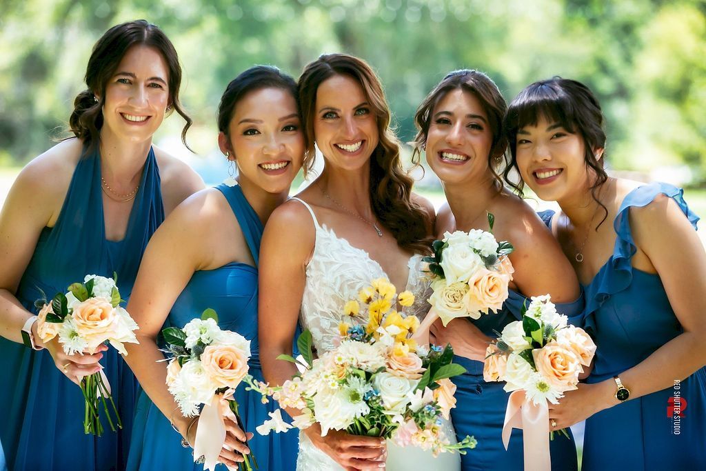 Five smiling bridesmaids in blue dresses holding bouquets outdoors at a wedding