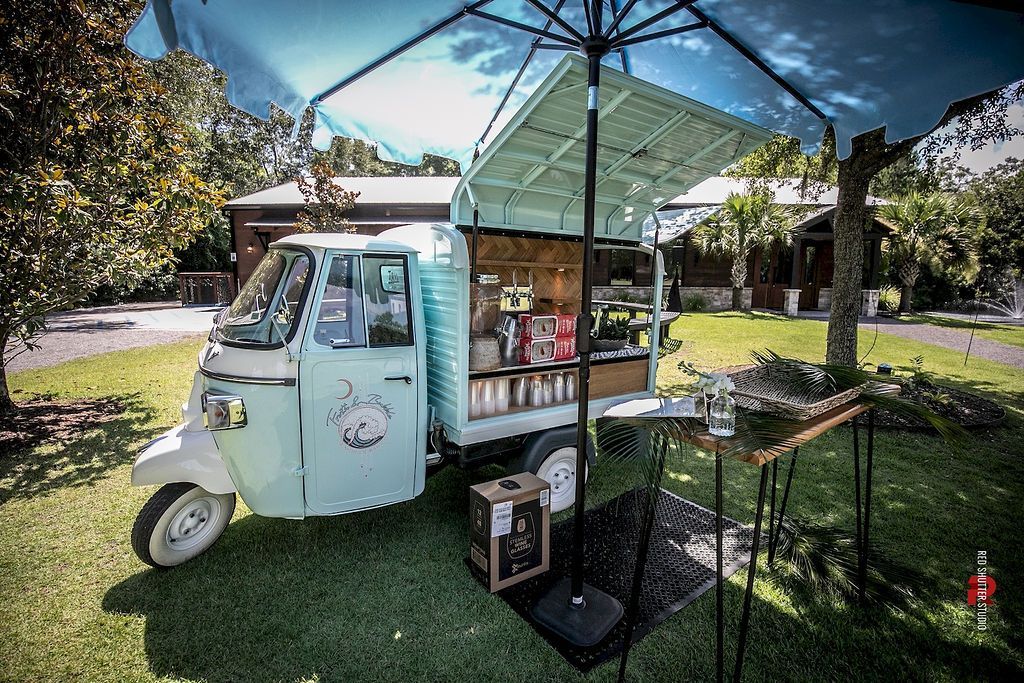 Pastel food truck under a blue umbrella in a sunny park, serving drinks and snacks from a small stand