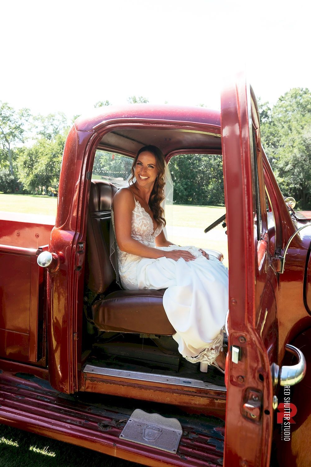 Woman seated in a vintage red truck with the door open, wearing a white dress and smiling outdoors