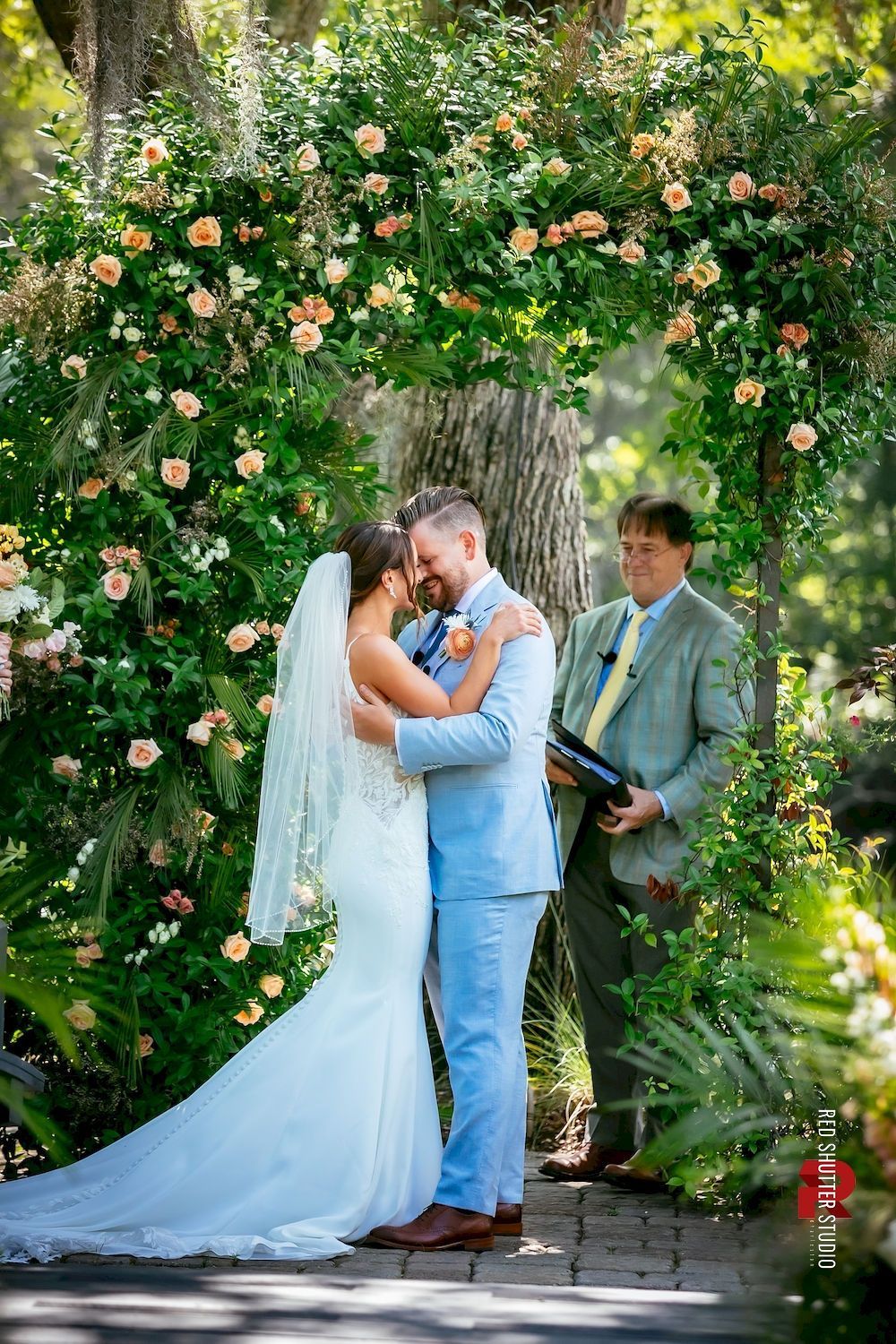 Wedding couple kissing beneath a flower-covered arch while an officiant stands nearby in a garden.