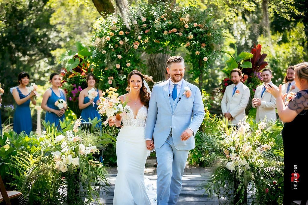Bride and groom walk down an outdoor floral aisle as guests applaud and take photos