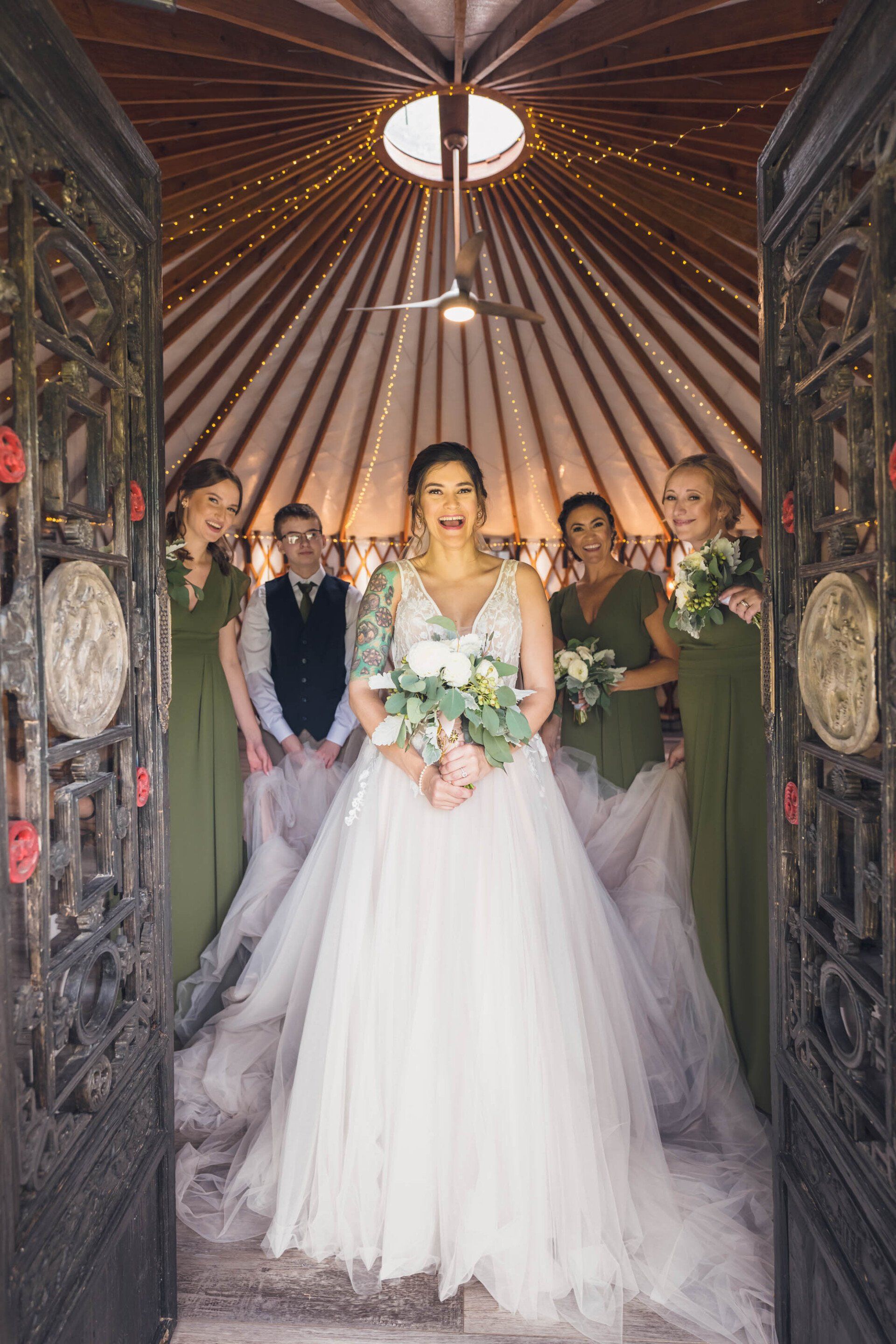 Bride in white gown holding bouquet, standing at an ornate doorway with wedding guests behind her.