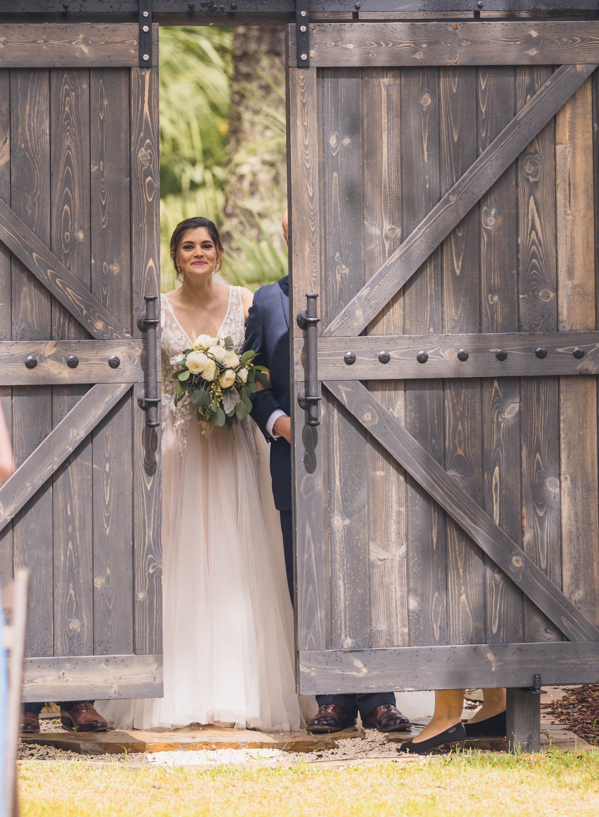 Bride in a white gown holding a bouquet, standing in a rustic barn doorway with wooden doors open.