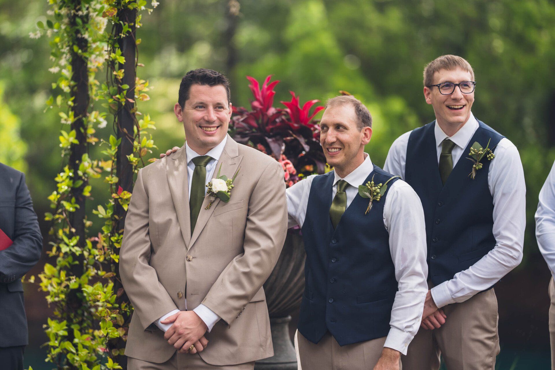 Three men in suits smiling outdoors at a wedding, with greenery and flowers behind them