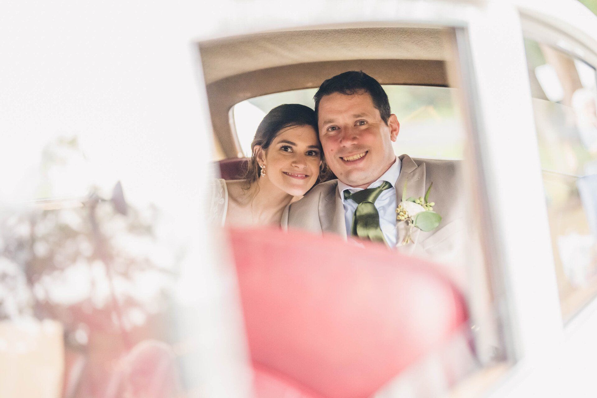 Smiling couple posing in a white car, seen through a mirror with soft pink foreground blur