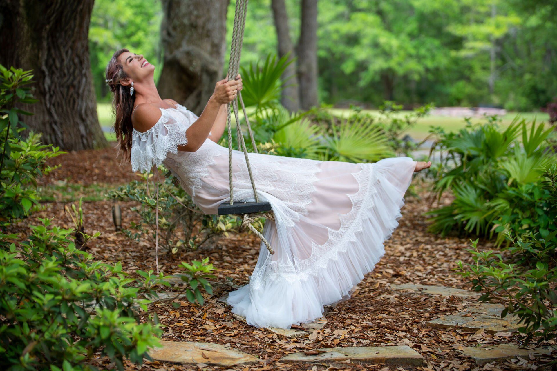Woman in a white dress swinging in a lush garden, surrounded by trees and greenery.