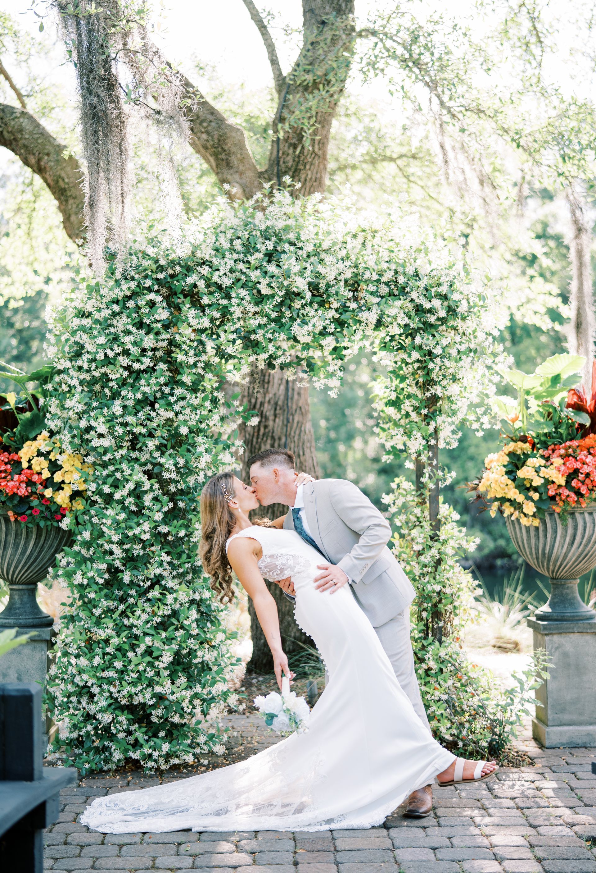Bride and groom kissing beneath a white floral arch in a garden setting