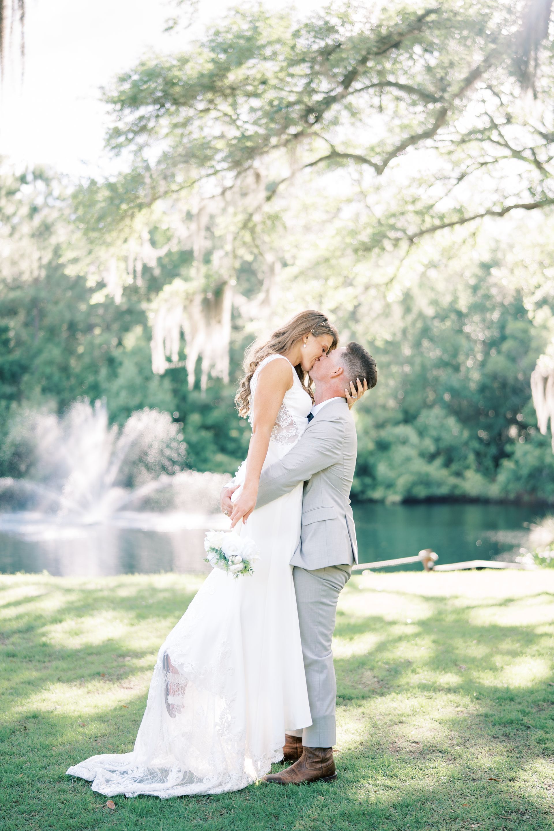 Couple in wedding attire embracing outdoors beside a fountain under a flowering tree