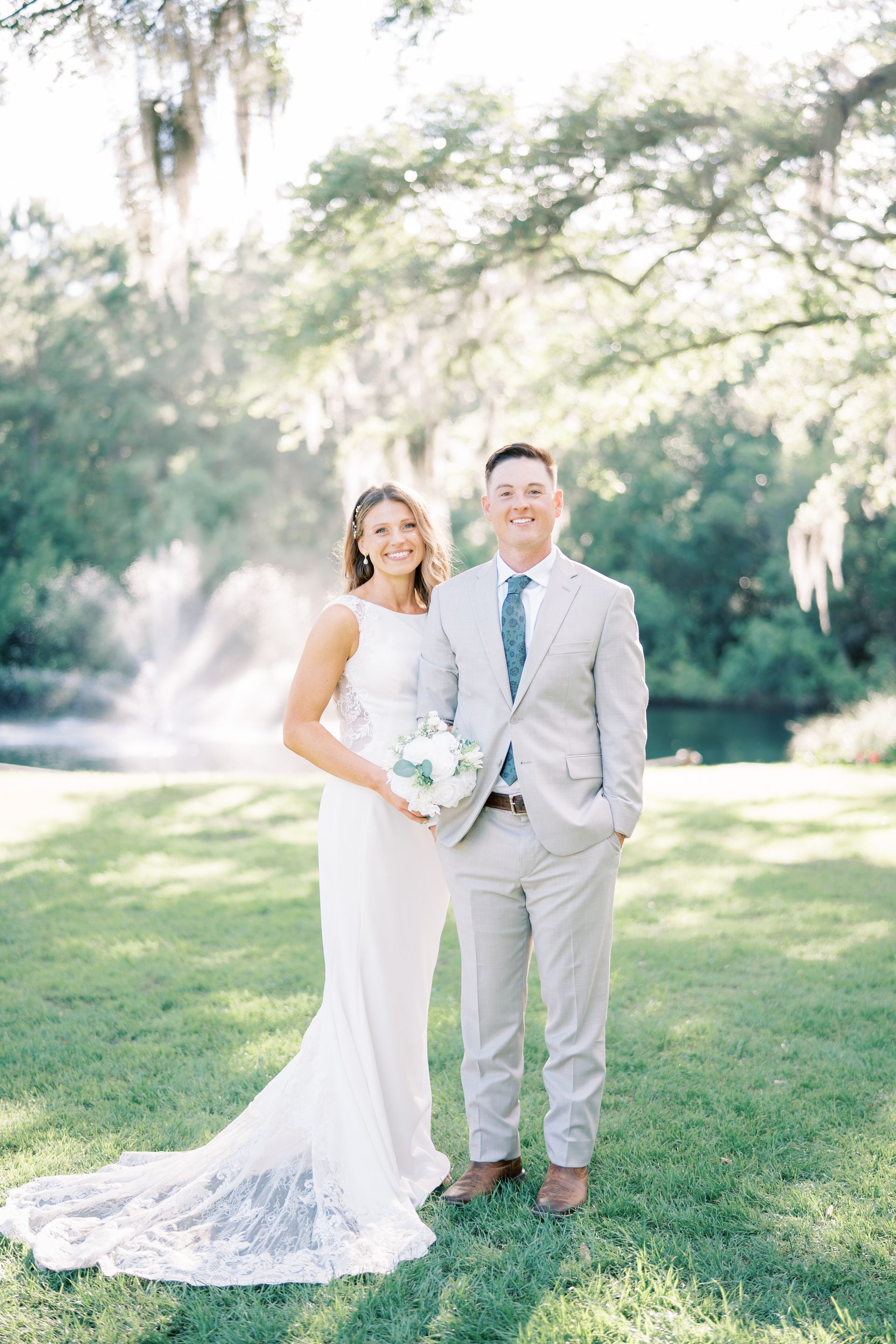 Bride and groom standing on a sunlit lawn, holding a bouquet in a bright outdoor setting