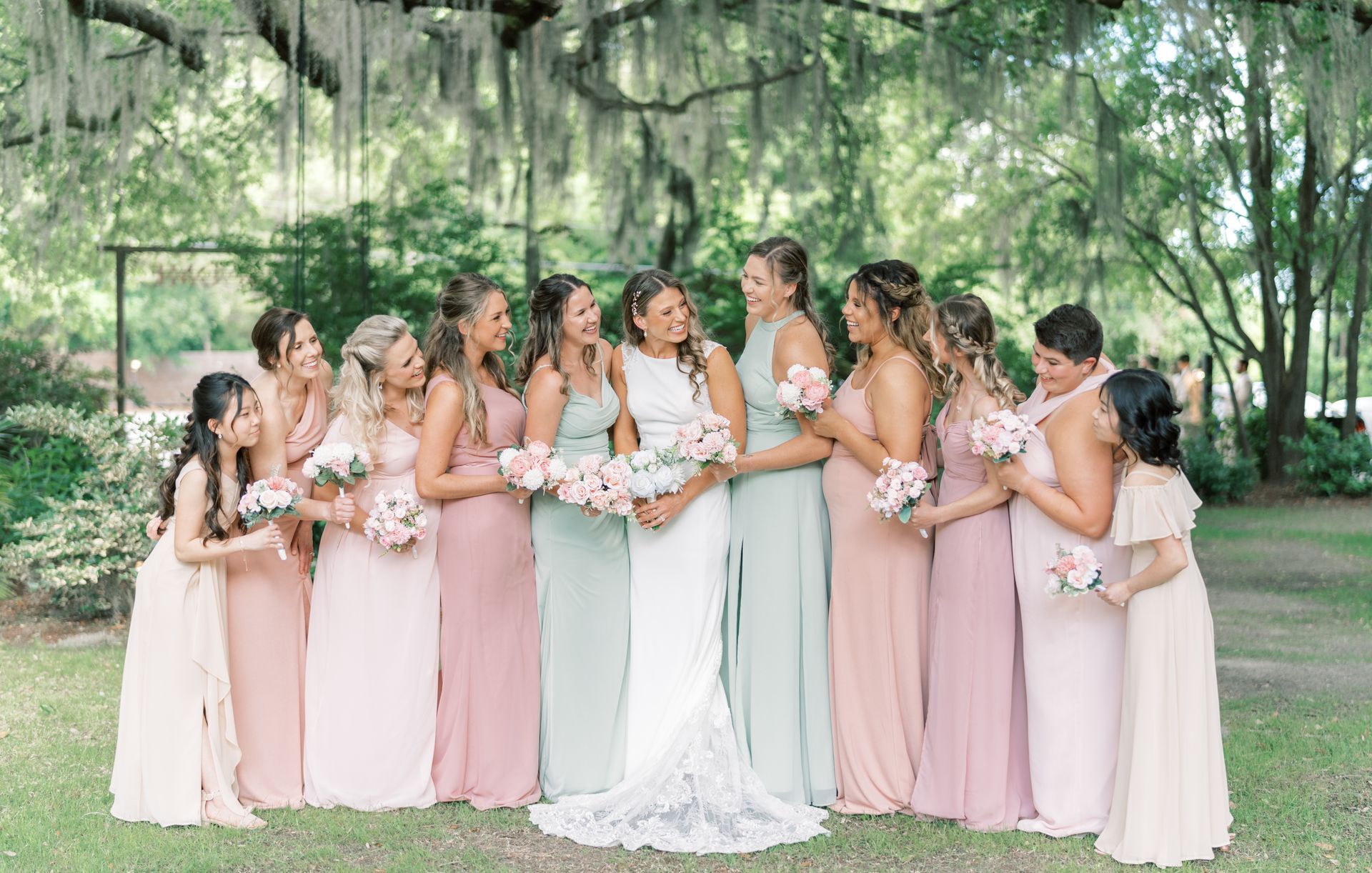 Bridal party in blush dresses and a white gown holding bouquets in a wooded outdoor setting
