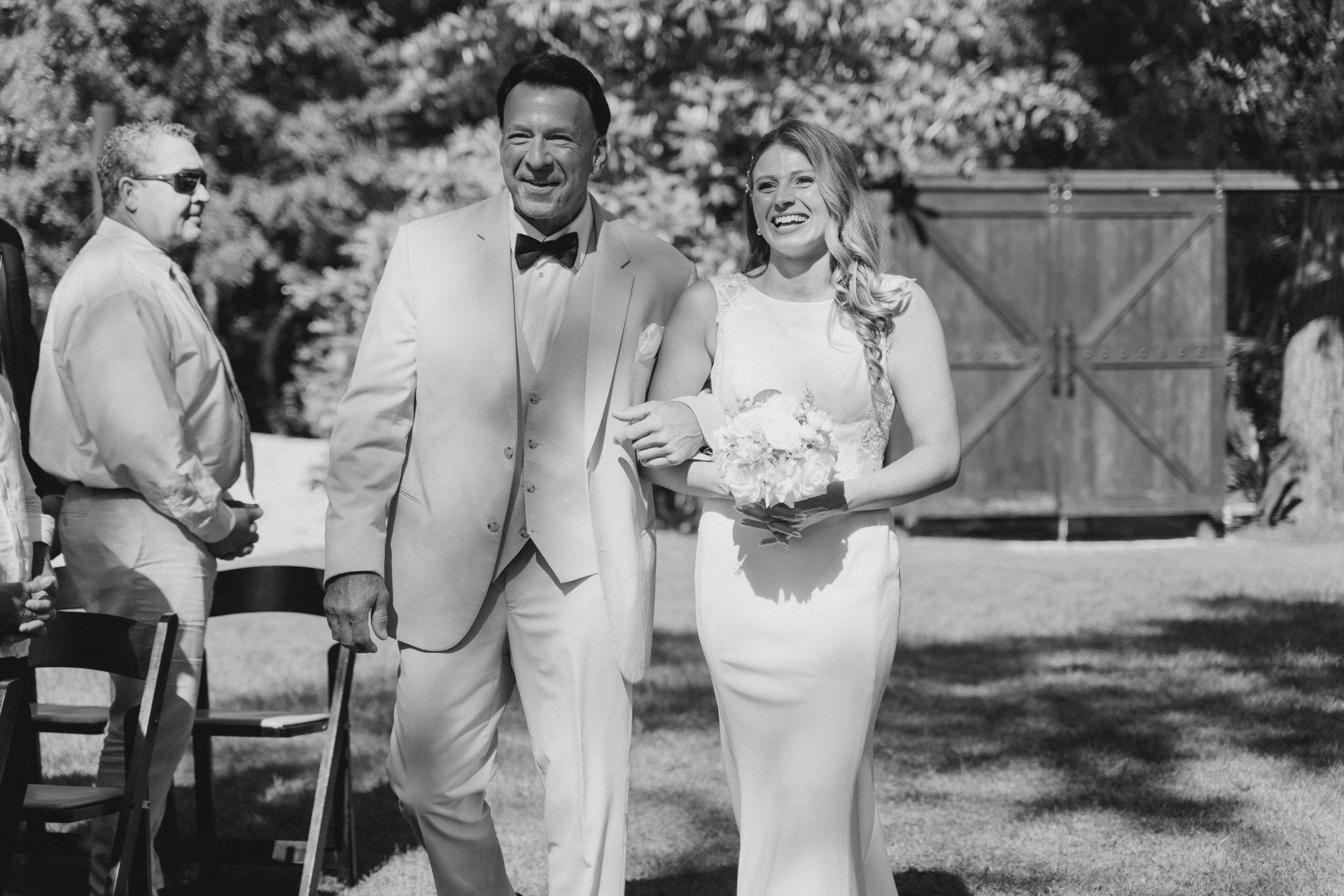 Three people in formal attire outdoors, with a groom and bride smiling near a wooden gate