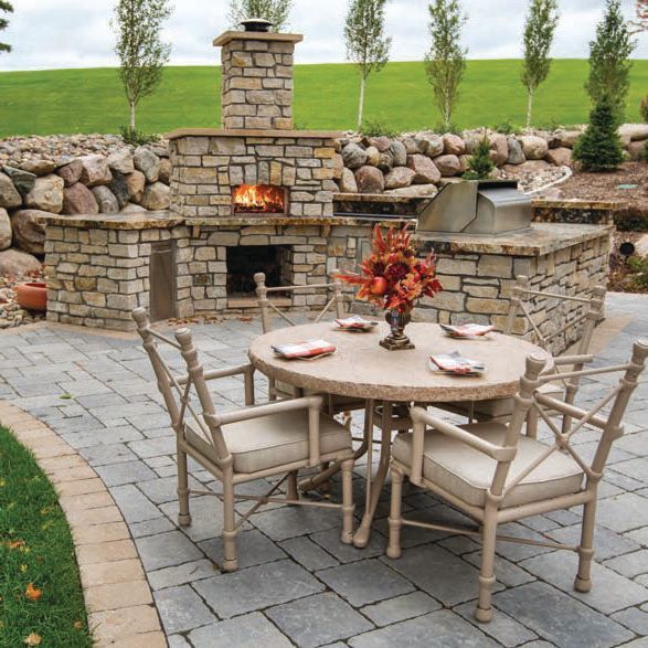 Stone patio with round table and chairs in front of an outdoor fireplace and rock wall