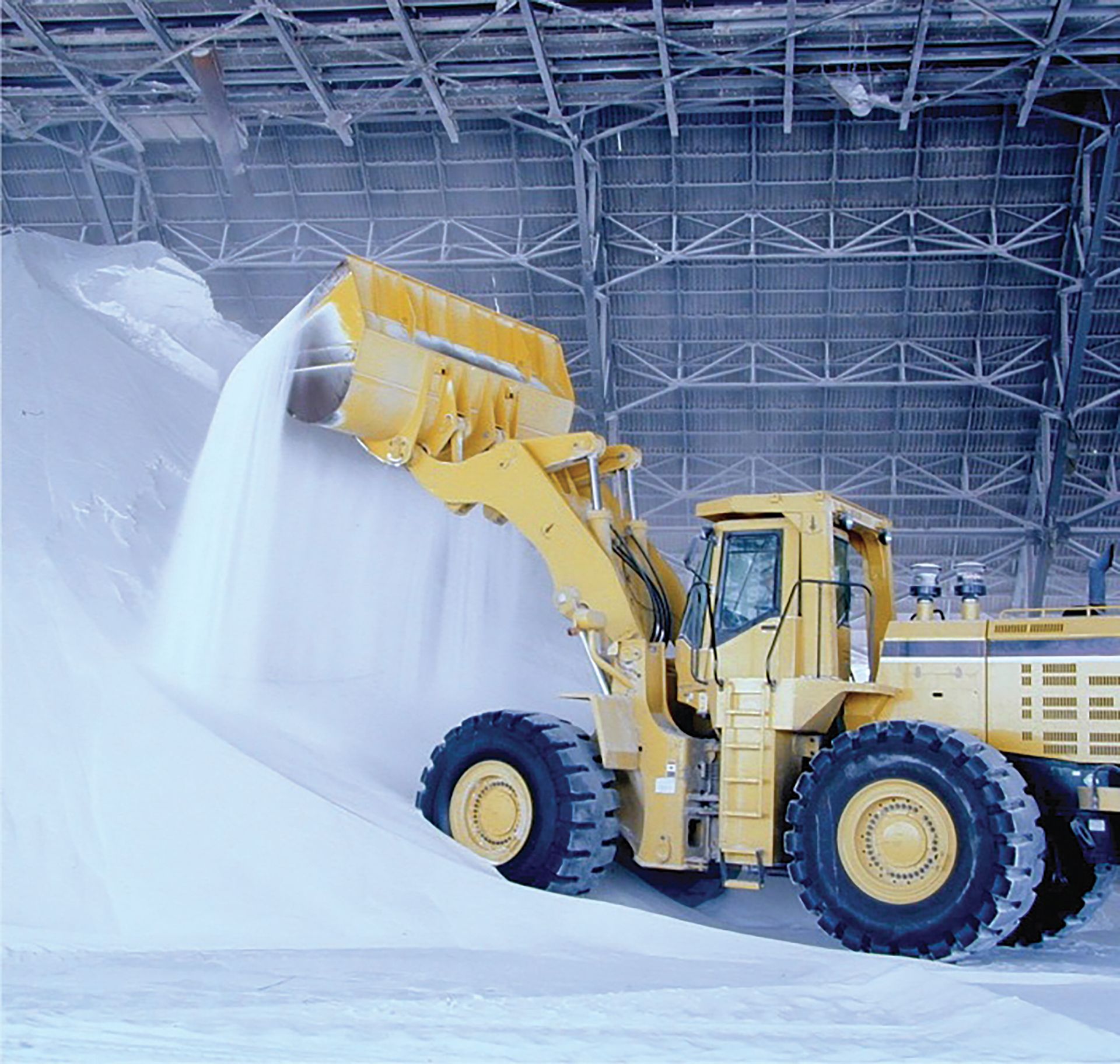 Yellow front loader dumping white material in a large indoor industrial warehouse