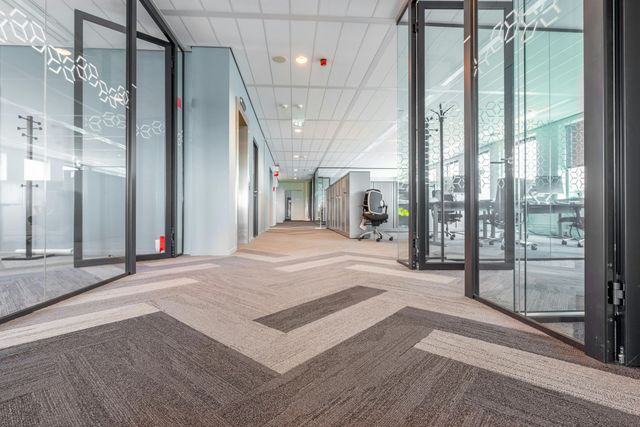 Modern office hallway with glass walls, carpeted floor, and open doors leading to workspaces.