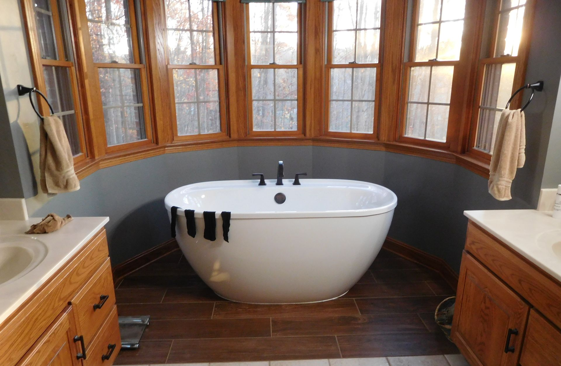 Bathroom with a white freestanding tub set in a bay window alcove between two wooden vanities.