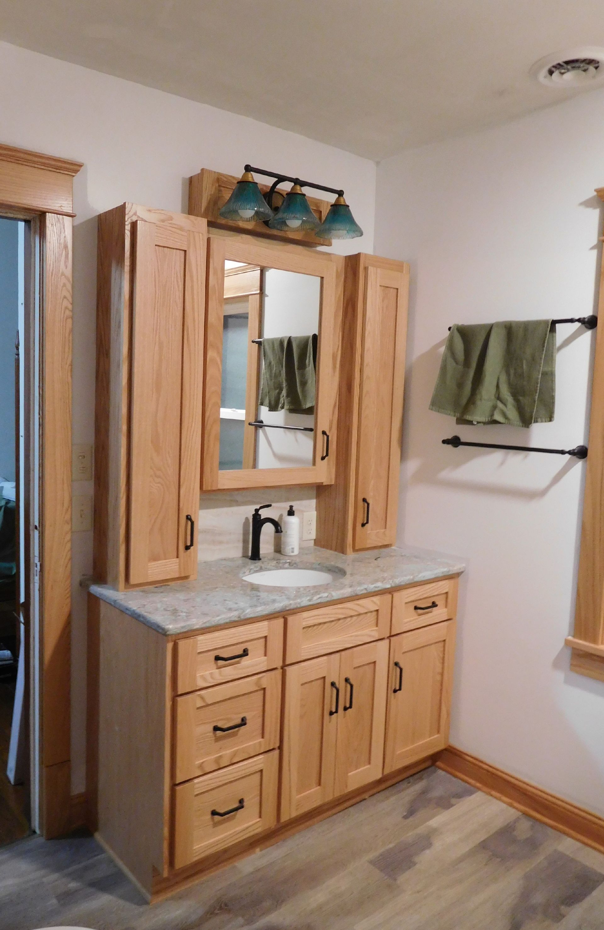 Bathroom with light-wood cabinets, light-colored countertops, a mirror, and a black towel rack.