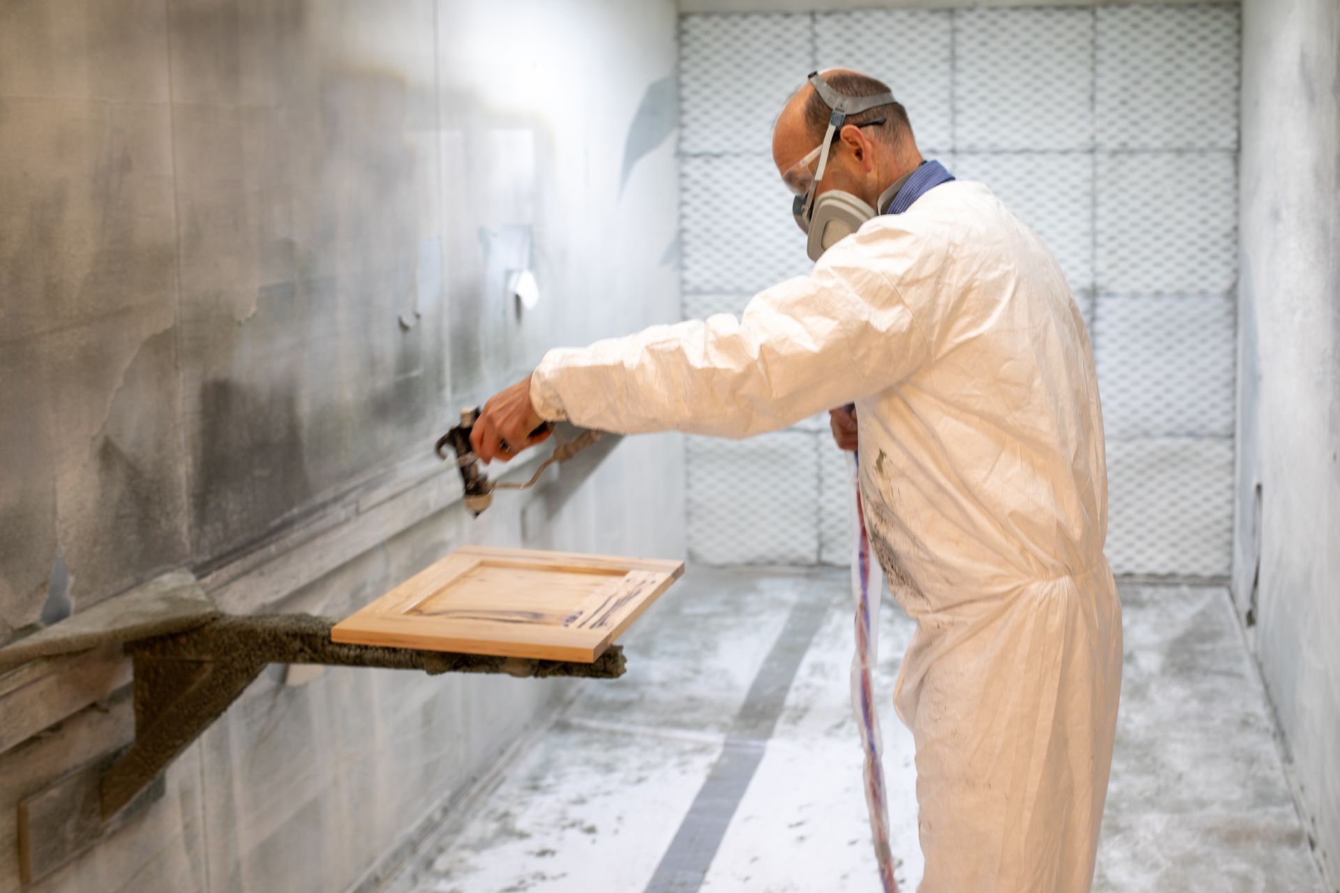Man in protective suit spray-painting a wooden cabinet door inside a paint booth.