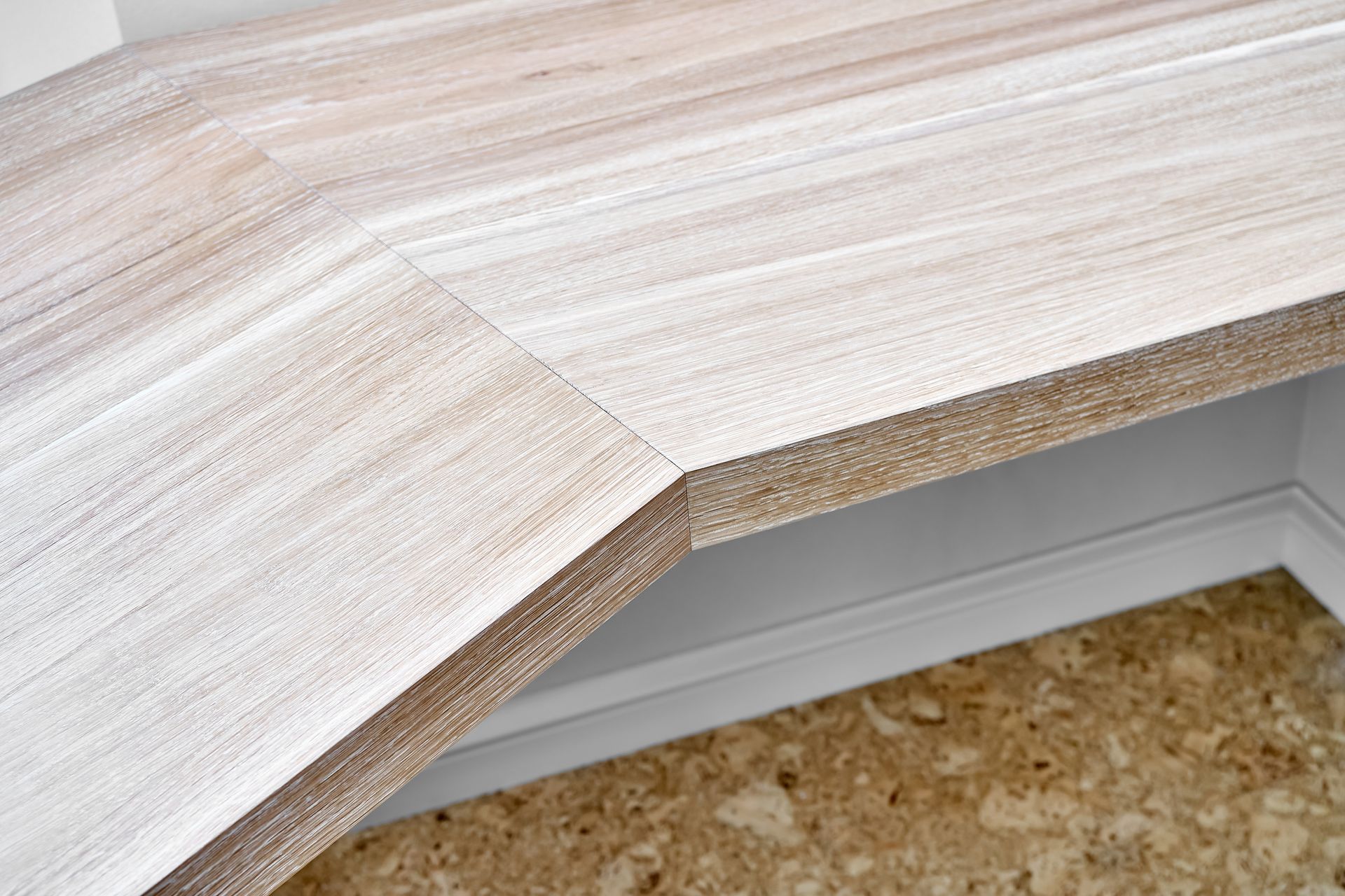 Close-up of a light-colored countertop with an angled edge, set above white trim and a mottled, beige wall.